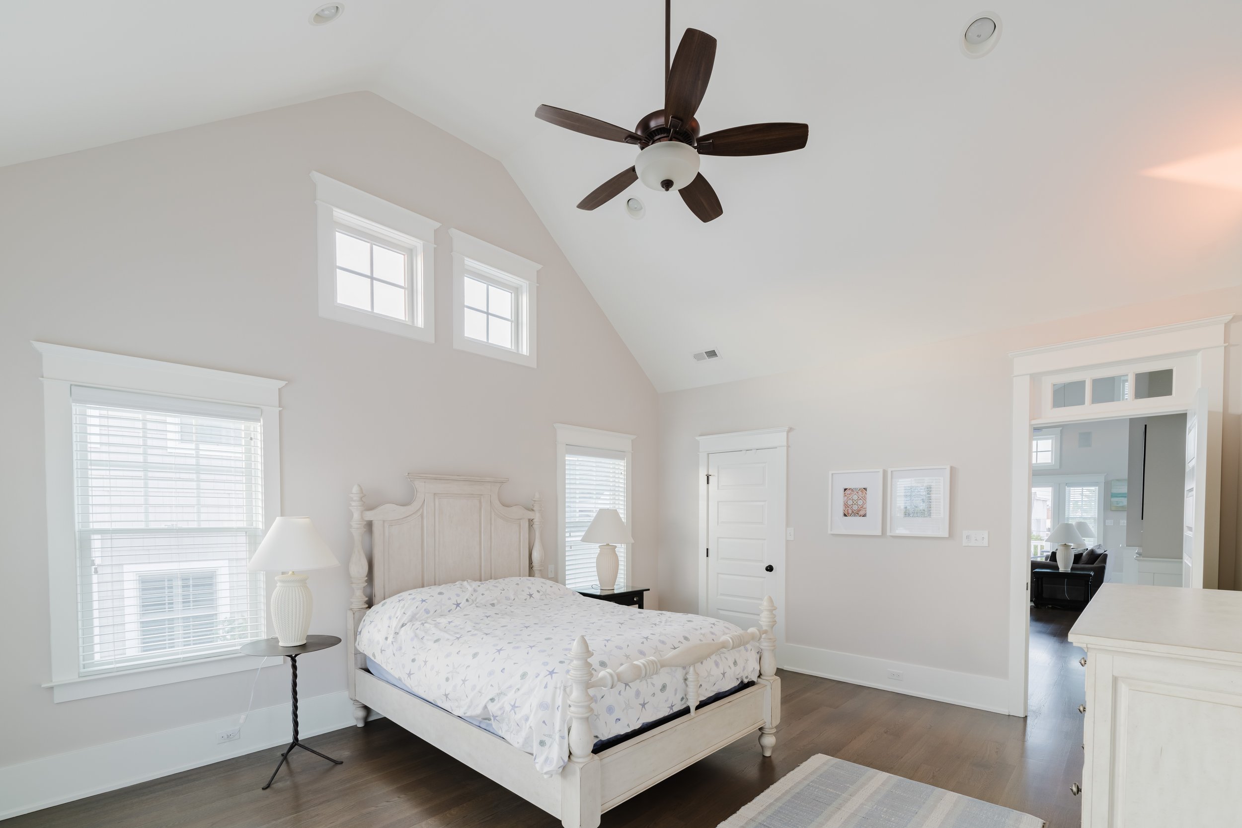 Elegant bedroom with neutral color palette featuring a wooden bed with a patterned bedspread, bedside lamps, large windows, ceiling fan, and framed artwork on the wall.
