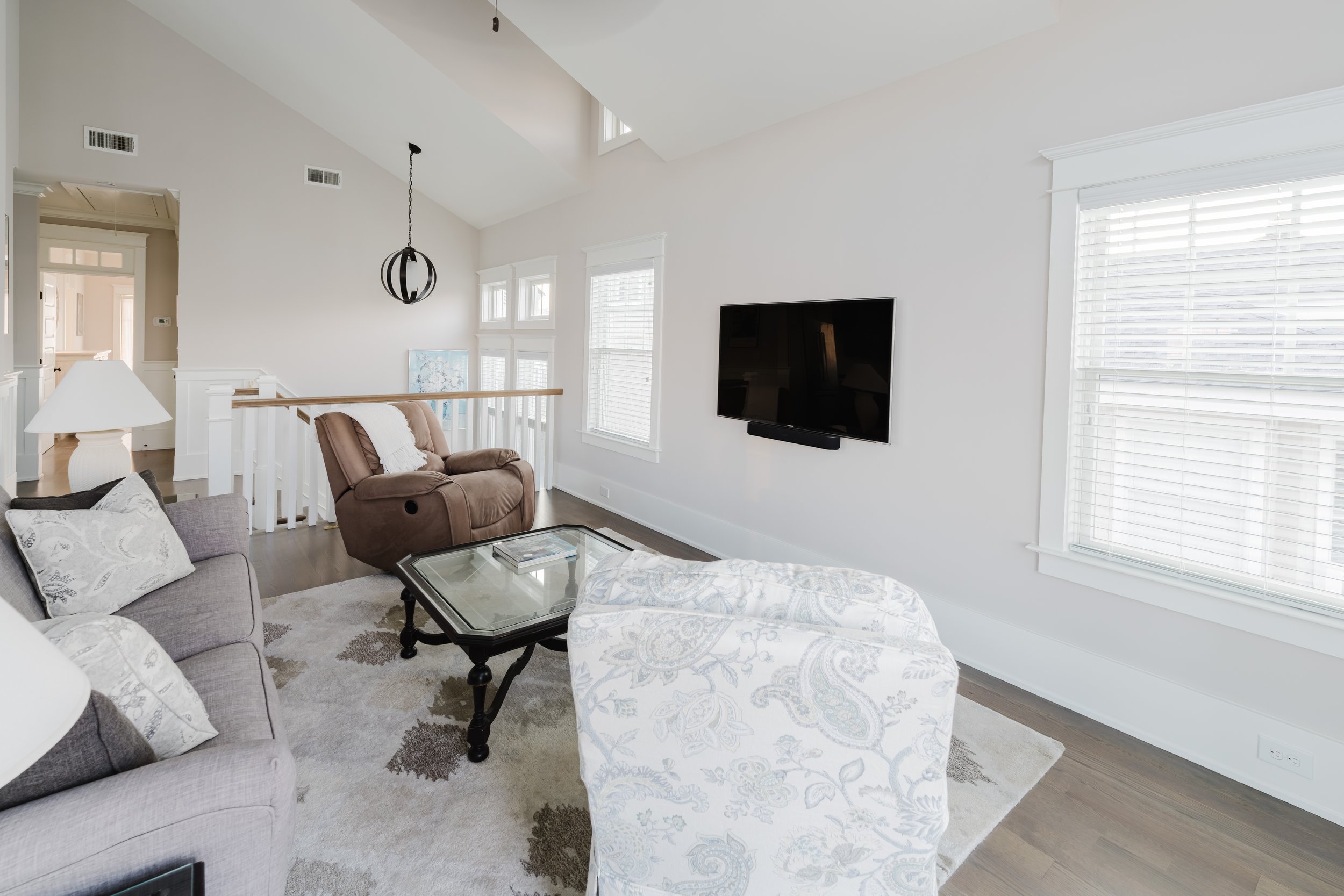 Modern living room with a gray sofa, patterned armchair, brown recliner, and glass coffee table on an area rug. A flat-screen TV is mounted on the wall, and large windows with white blinds provide natural light. A unique hanging light fixture is pres