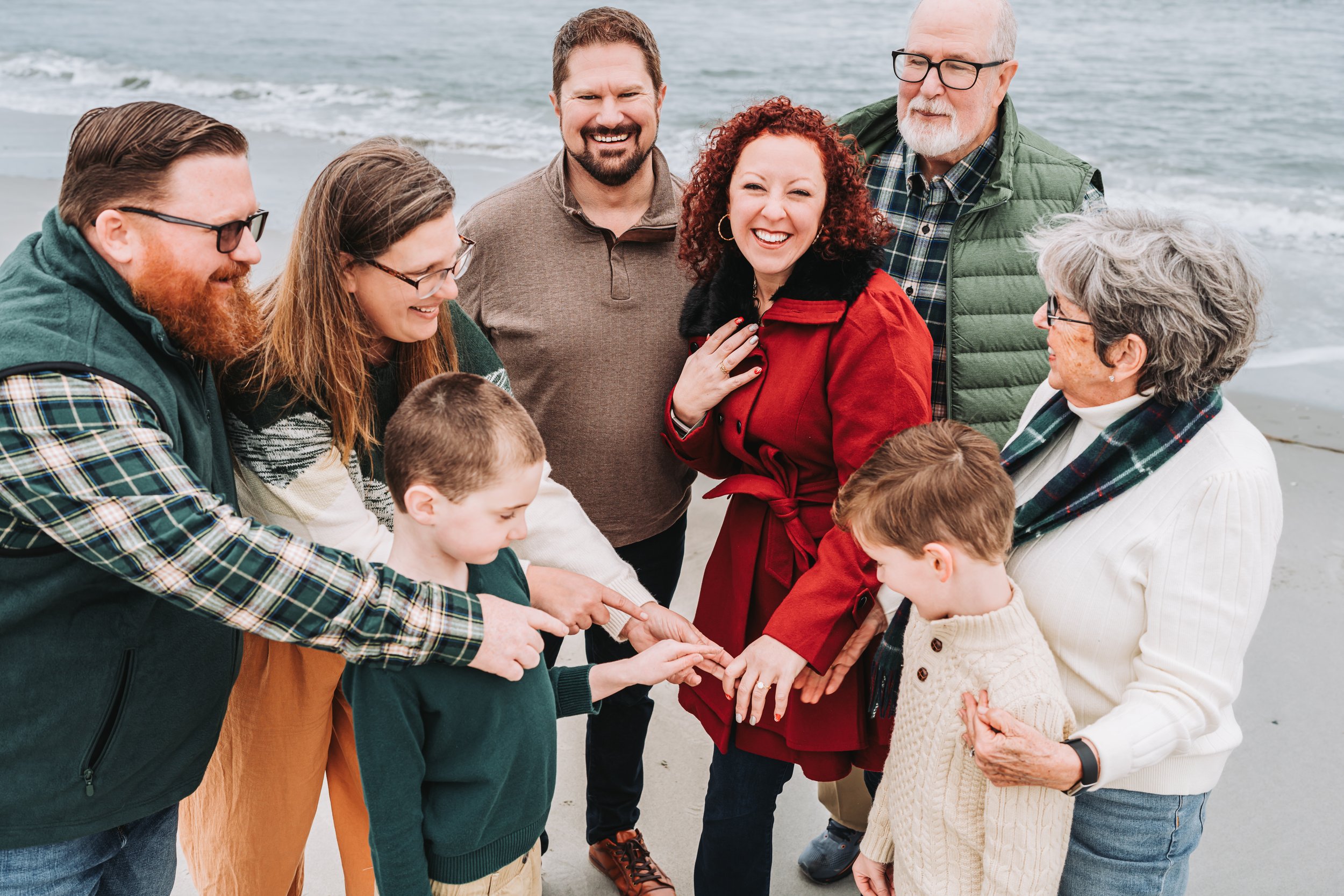 Group of people smiling on a beach, one woman in a red coat displaying a ring, surrounded by family members.