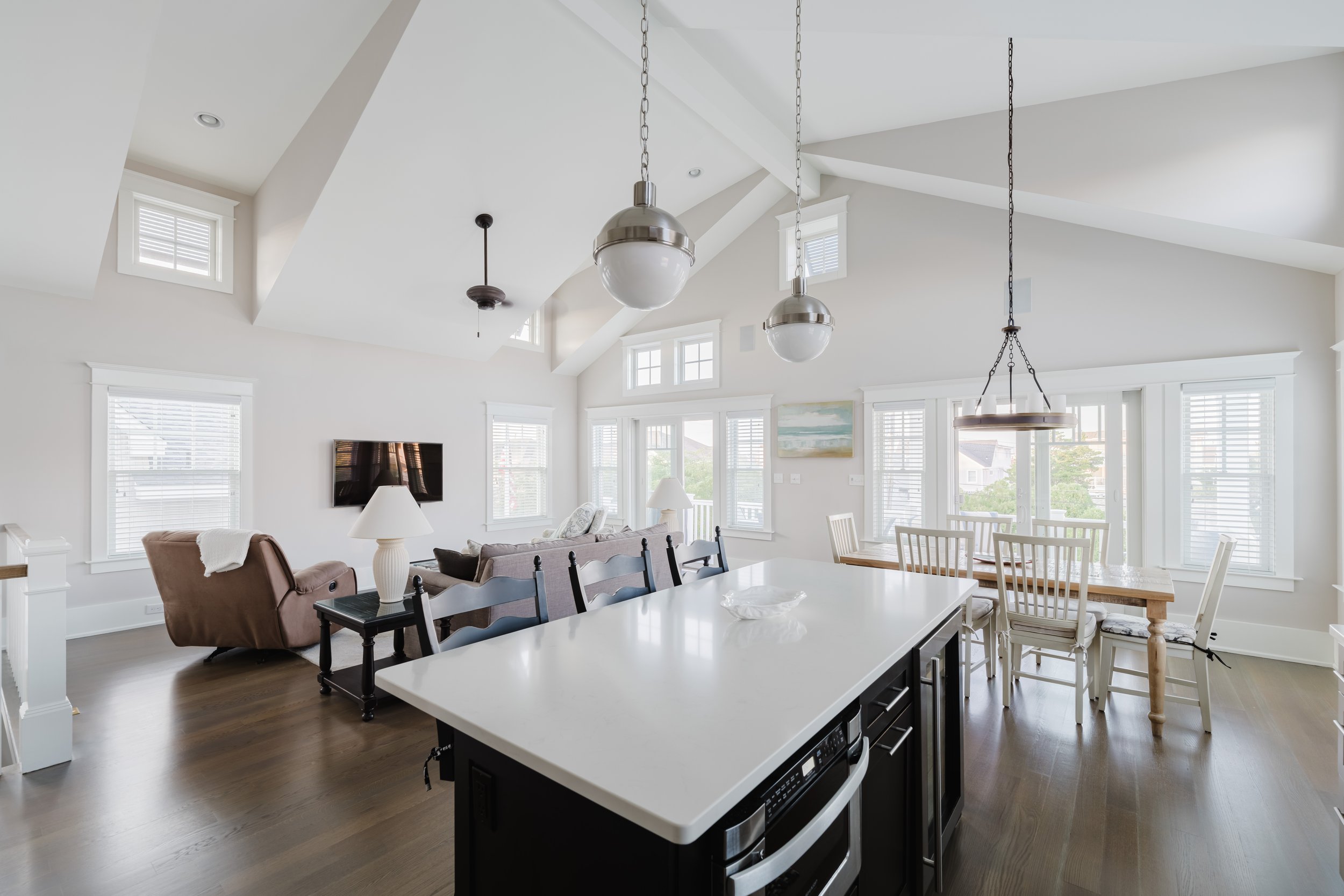 Open-concept modern living room and dining area with vaulted ceiling, featuring a kitchen island, hanging pendant lights, a dining table with chairs, and comfortable seating. Large windows enhance natural lighting.