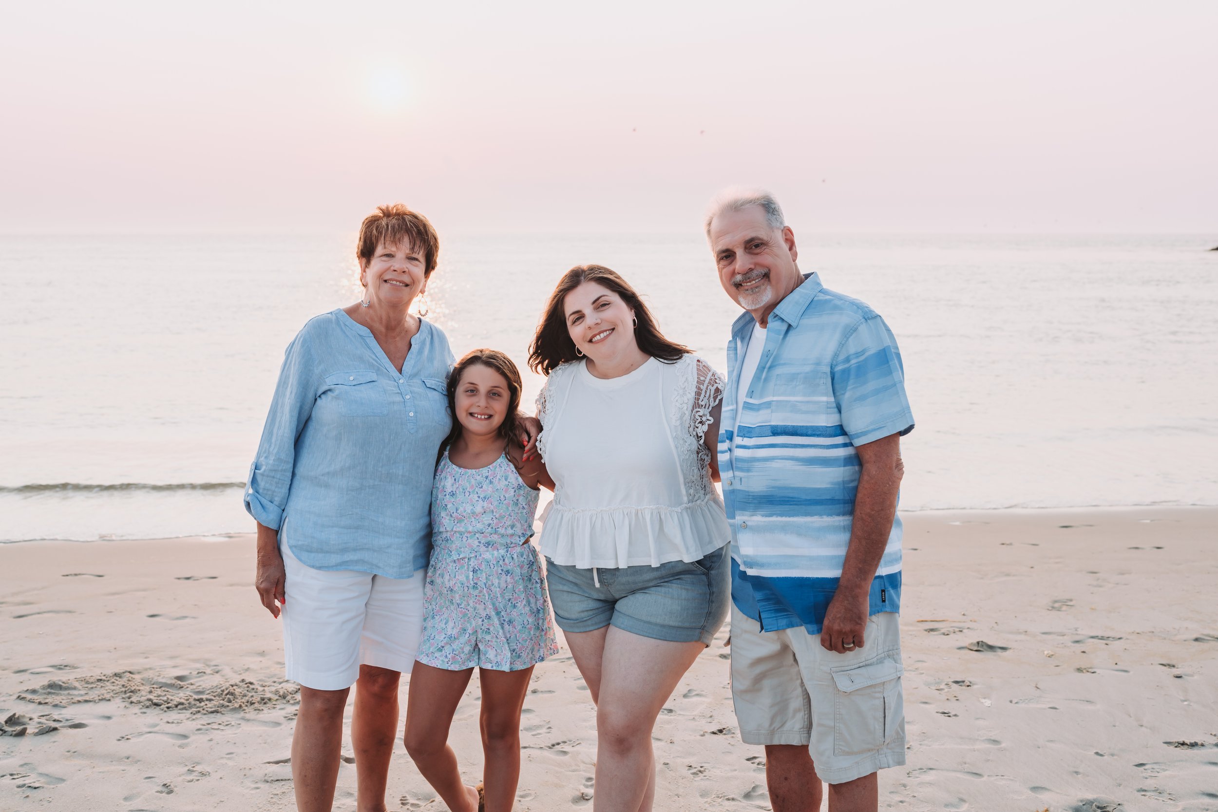 A family of four posing on a beach at sunset, with the ocean in the background. An elderly woman in a blue shirt stands next to a young girl in a floral dress. Beside them, a woman in a white top and shorts smiles, alongside an older man in a blue st