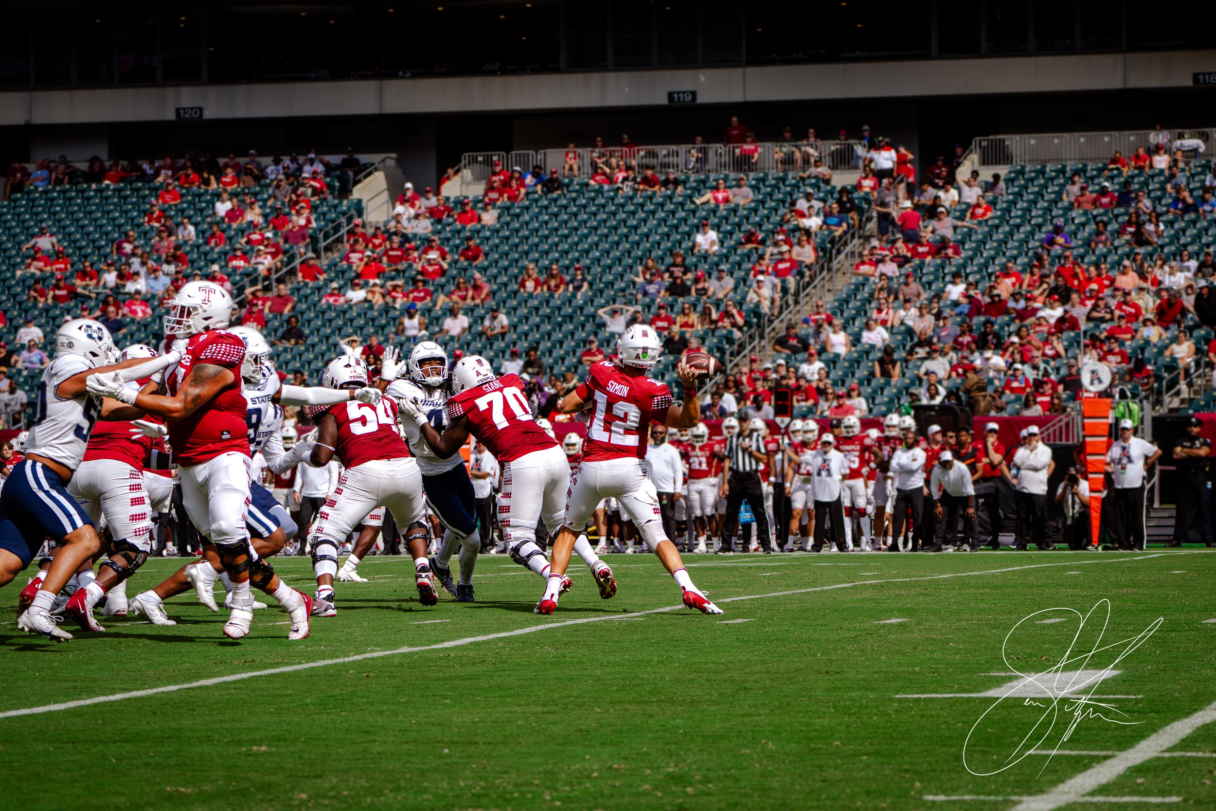 American football game with players in red uniforms, including a quarterback throwing the ball, defending against players in white and blue uniforms. The audience is visible in the stadium background.