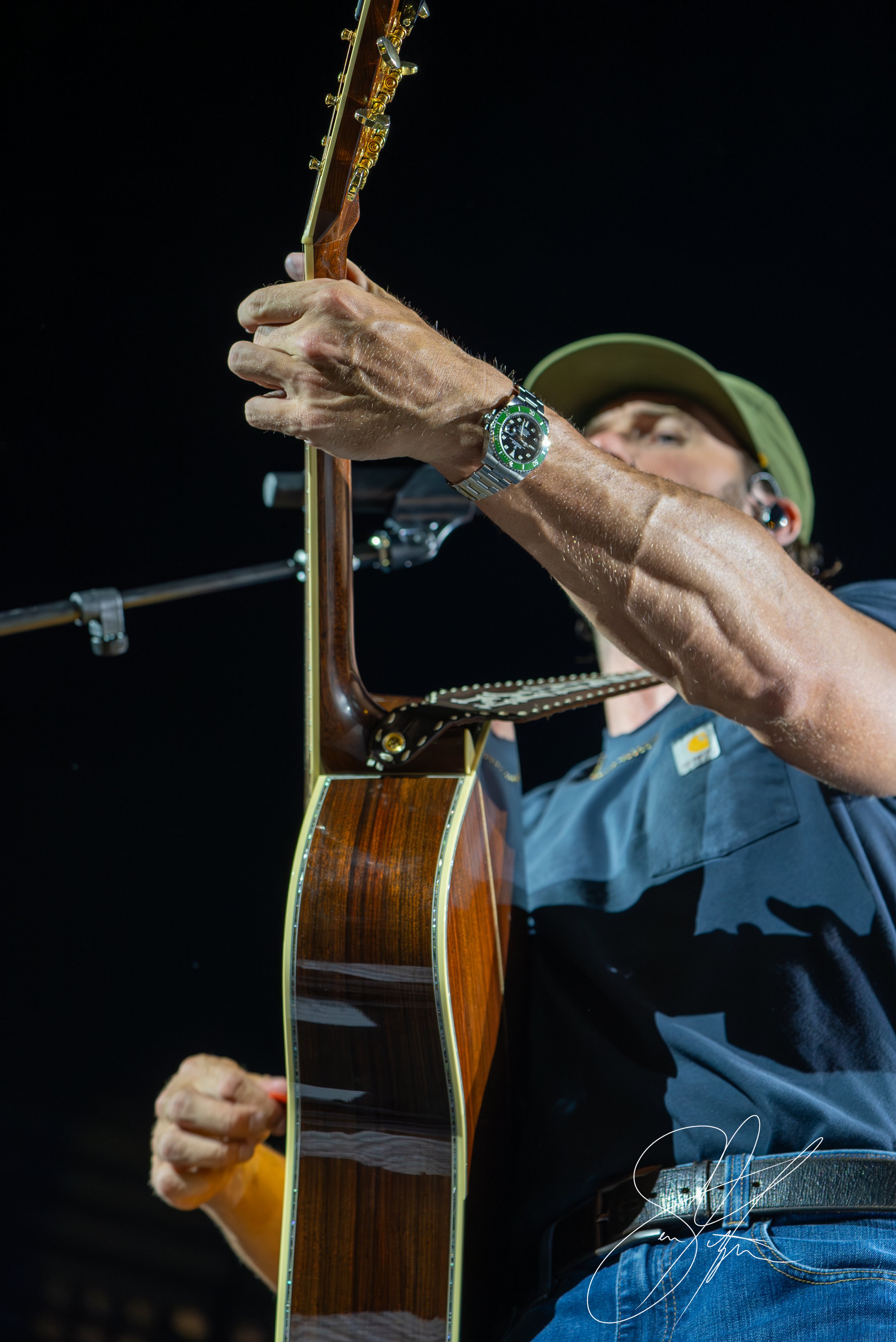 Close-up of a person's arm holding an acoustic guitar, wearing a wristwatch, with a microphone stand and dark background.