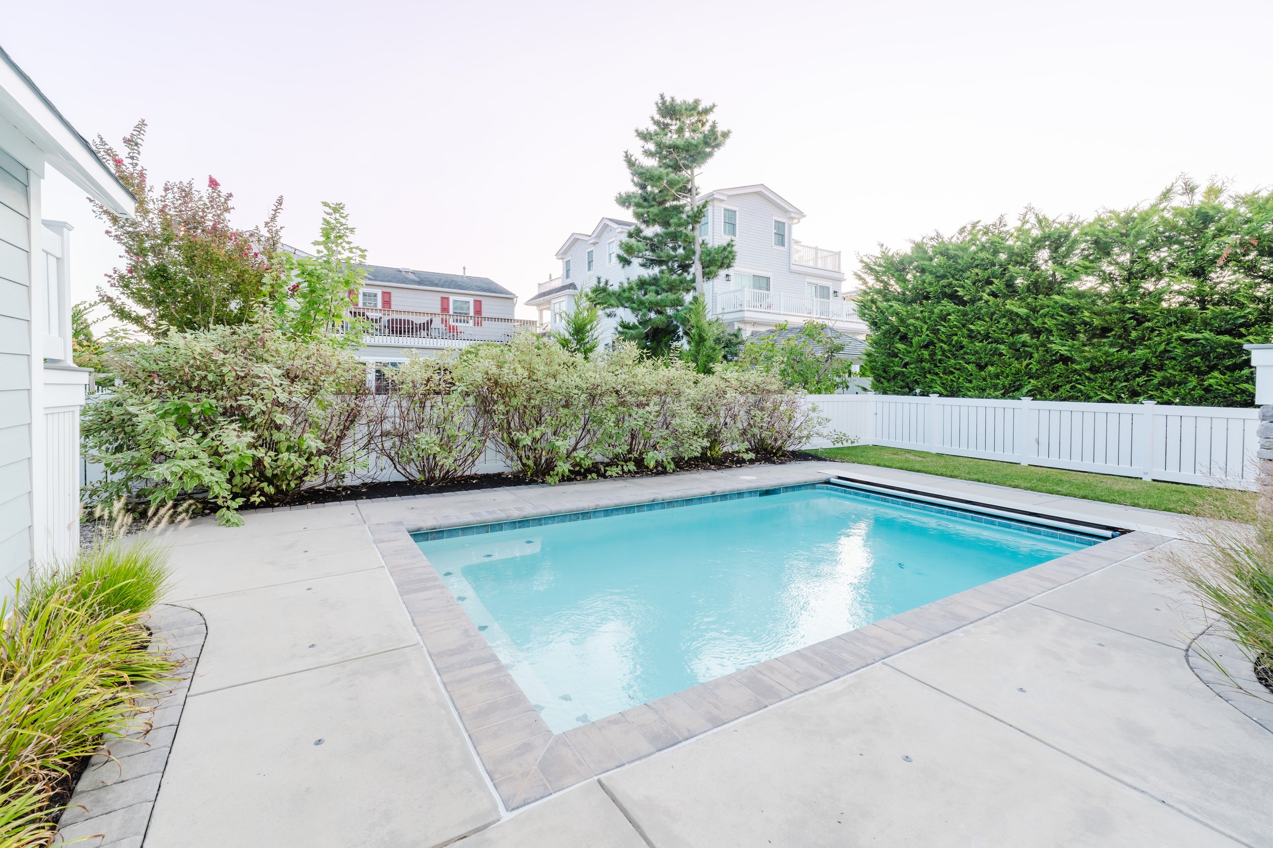 Backyard with a small rectangular swimming pool, surrounded by concrete pavement and lush green shrubs, with white fences and houses in the background.
