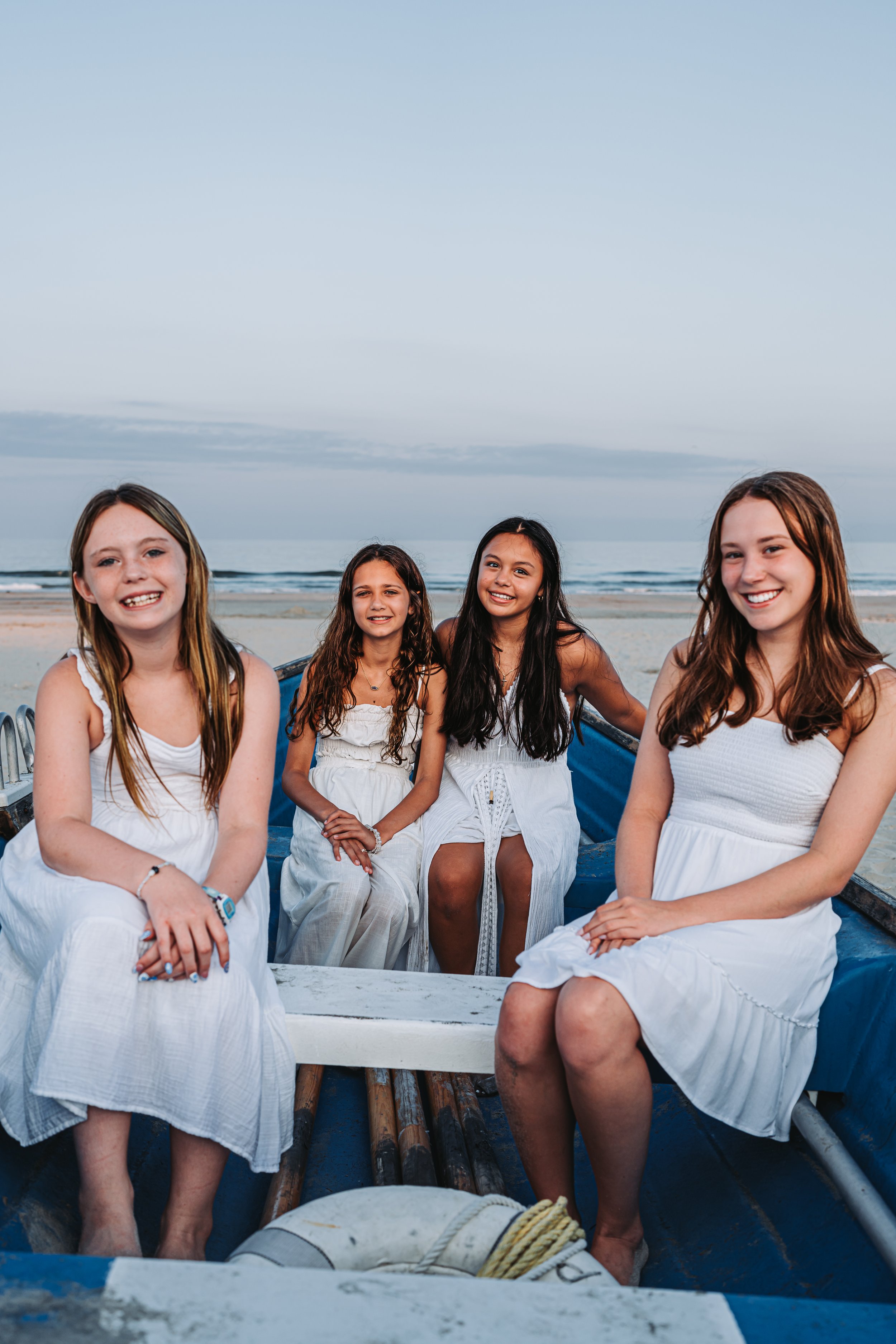 Four girls in white dresses sitting in a boat on a beach with ocean in the background.