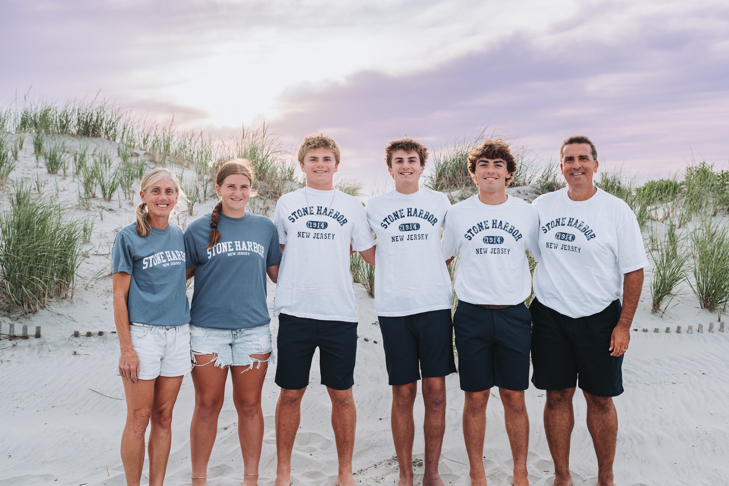 Family posing on a sandy beach wearing matching Stone Harbor New Jersey shirts.