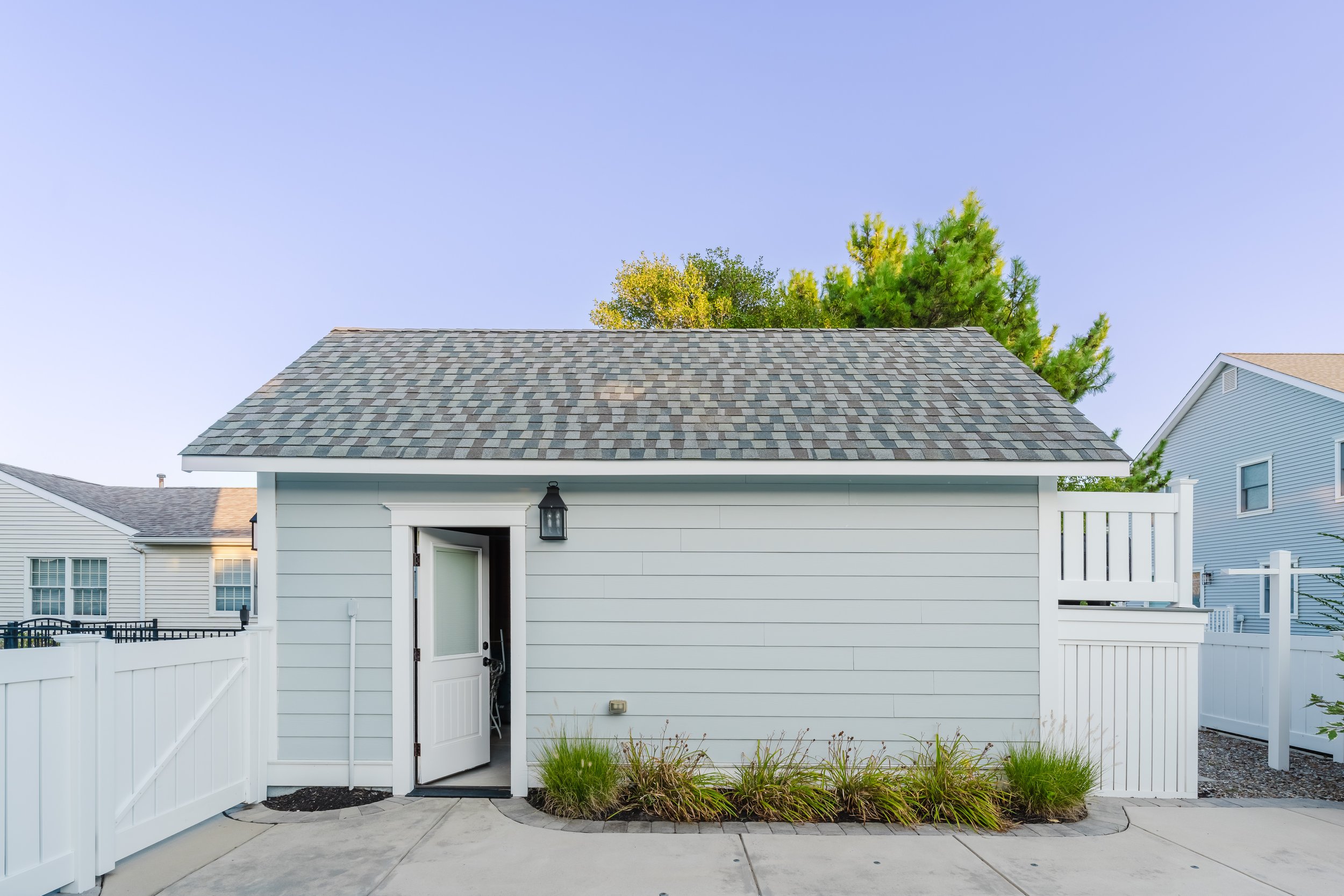 Small gray shed with shingle roof, open door, outdoor light fixture, white fence, and plants; adjacent houses visible.