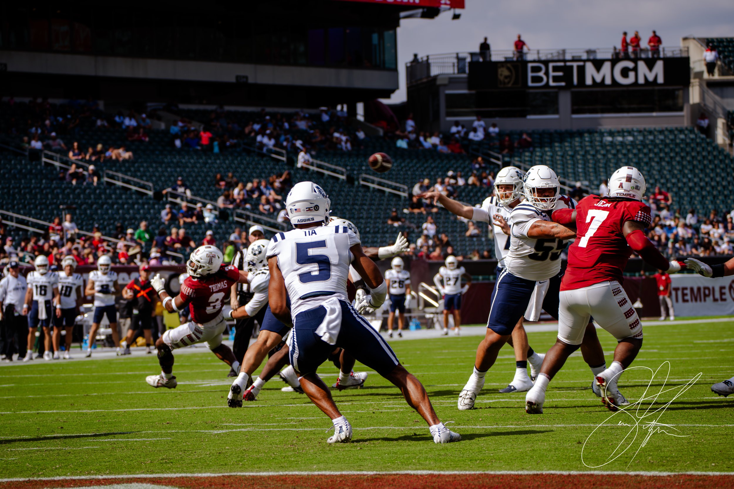 College football game with players in action on the field, wearing red and white jerseys, with a stadium crowd in the background.