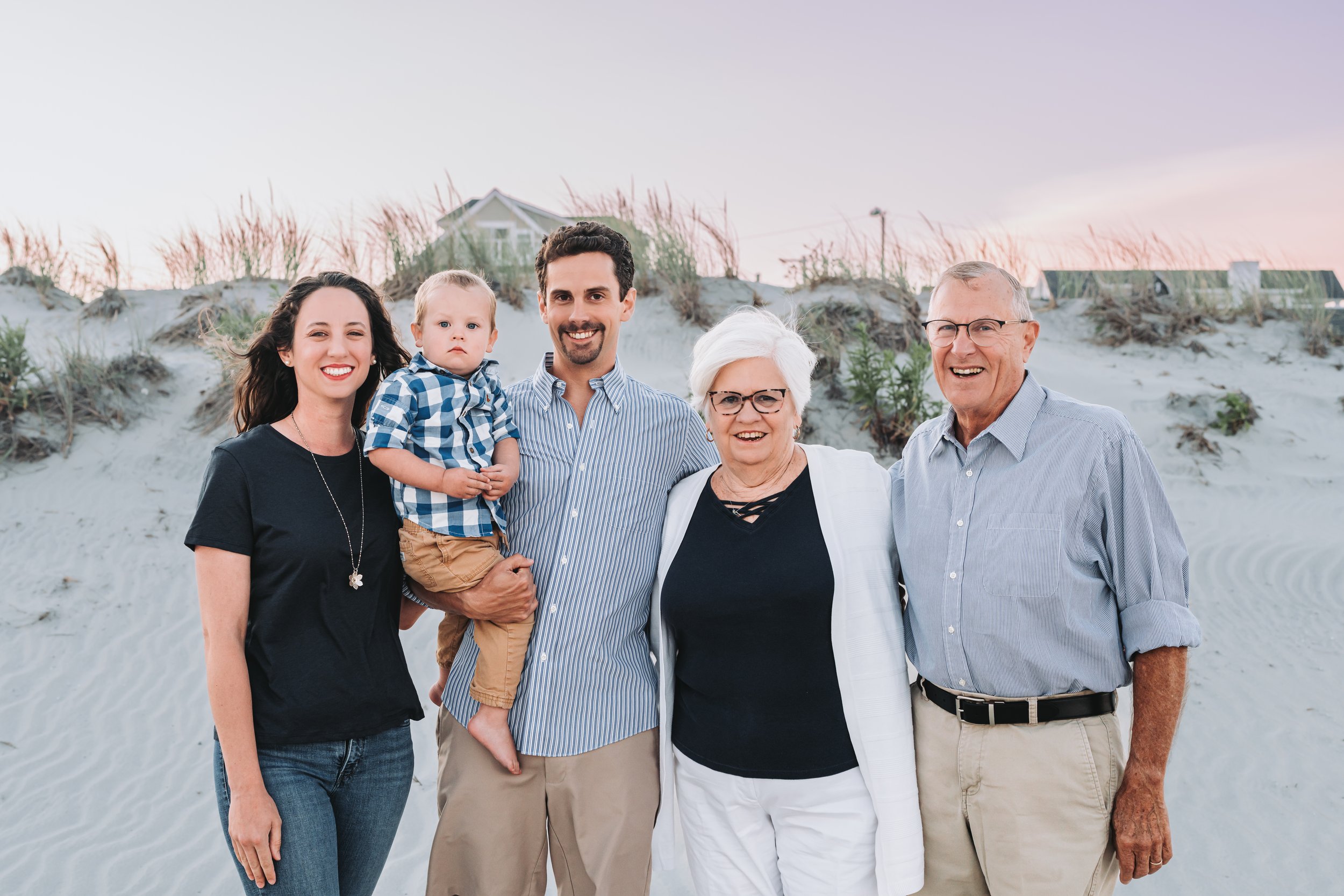 Family of five smiling on a sandy beach with grass and a house in the background during sunset.