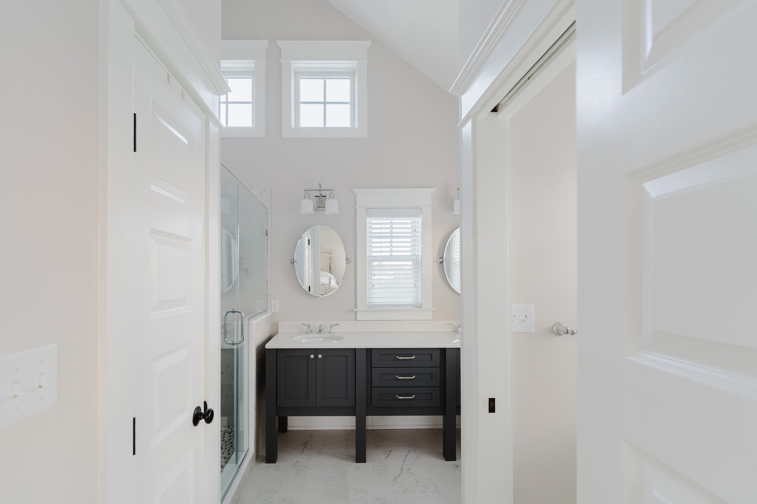 Modern bathroom with a glass shower, double vanity with dark cabinets, two oval mirrors, and large windows.