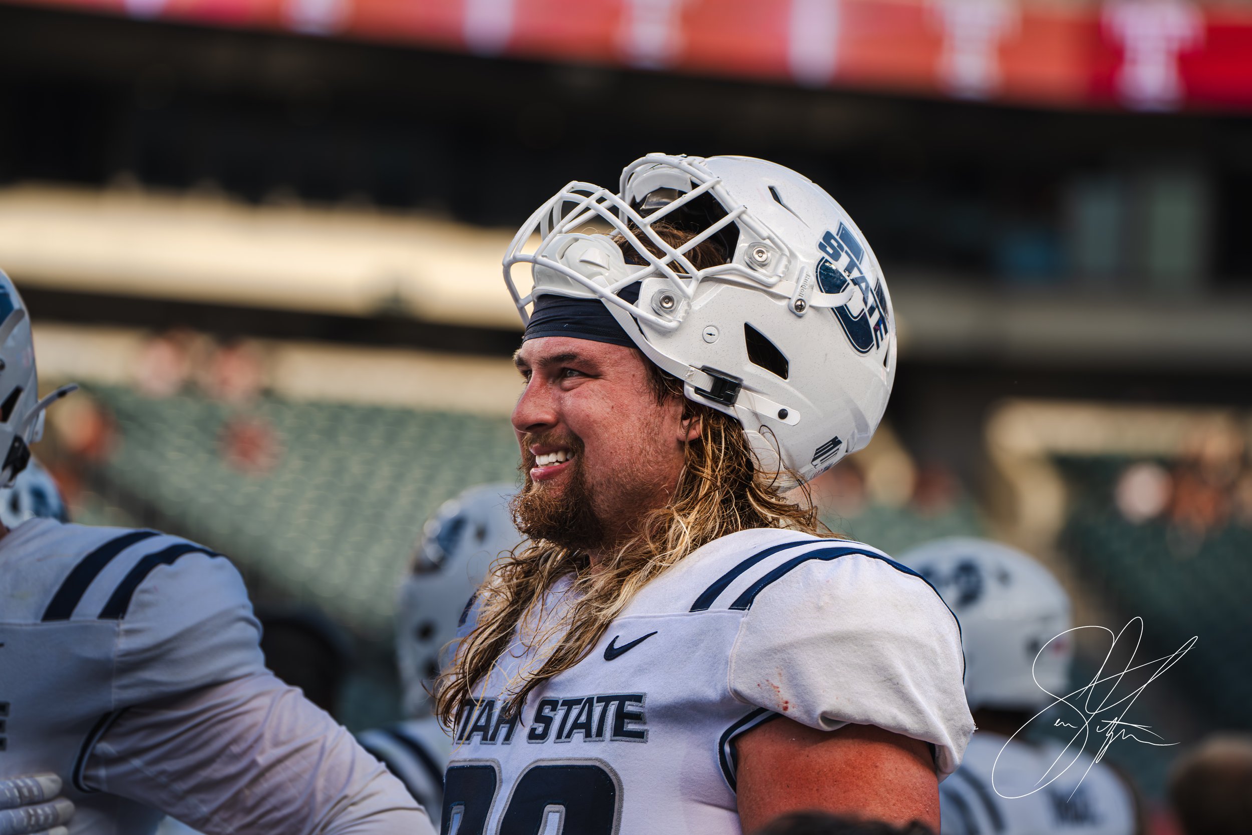 Football player wearing a white "Utah State" uniform and helmet, smiling on the field with stadium seats in the background.