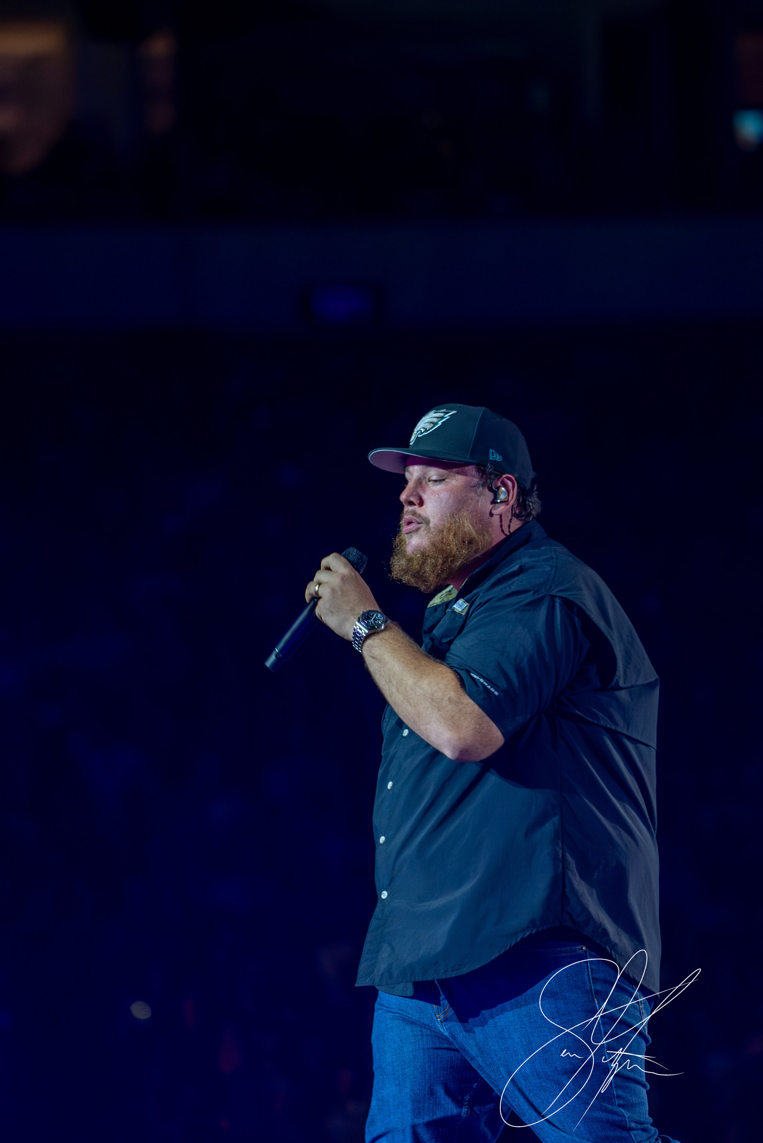A man wearing a dark cap and shirt holding a microphone on stage with dark background, performing or speaking.