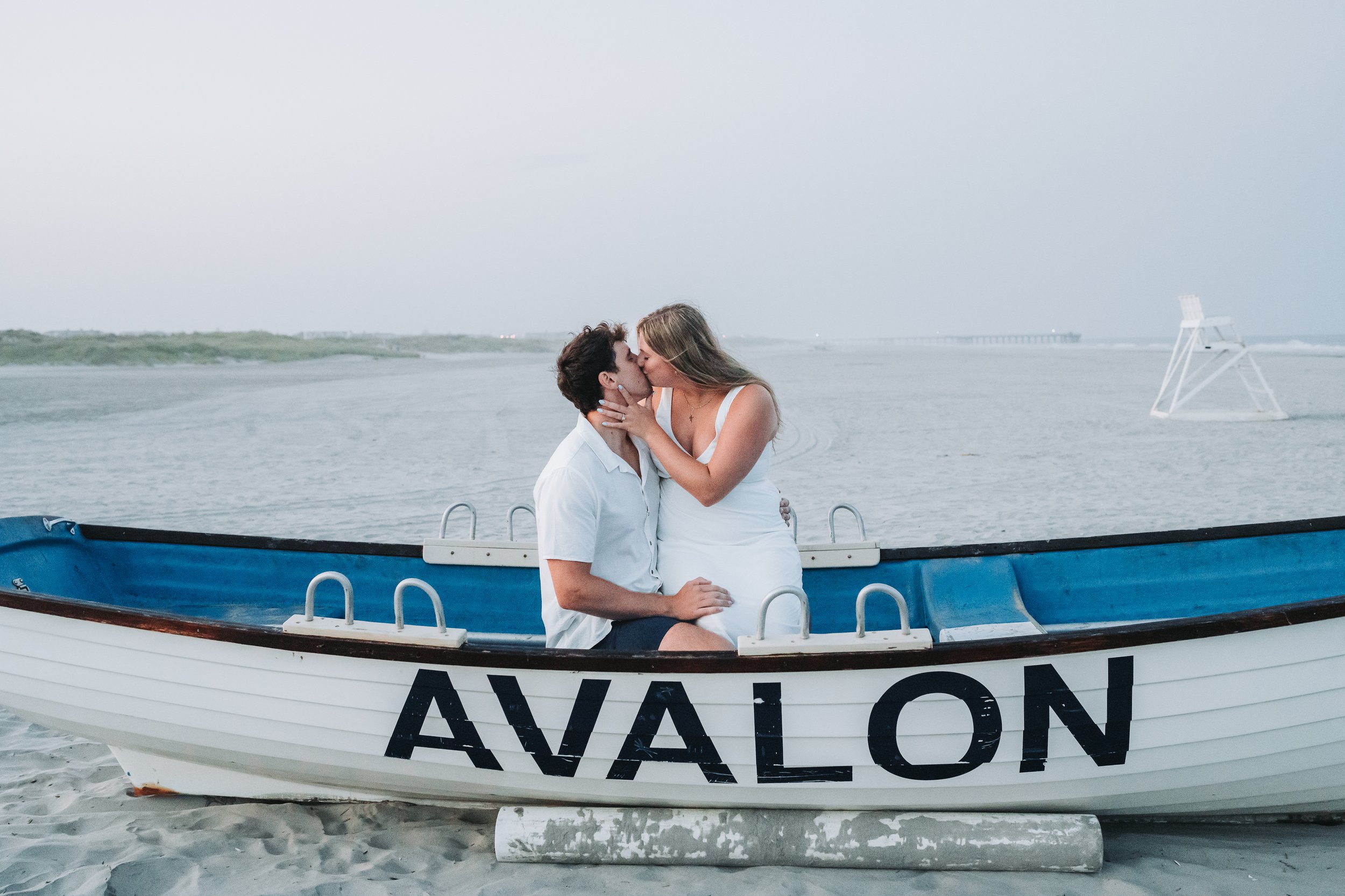 Couple kissing in a beached rowboat labeled 'AVALON' on a sandy shore with a distant view of the ocean and a lifeguard chair.
