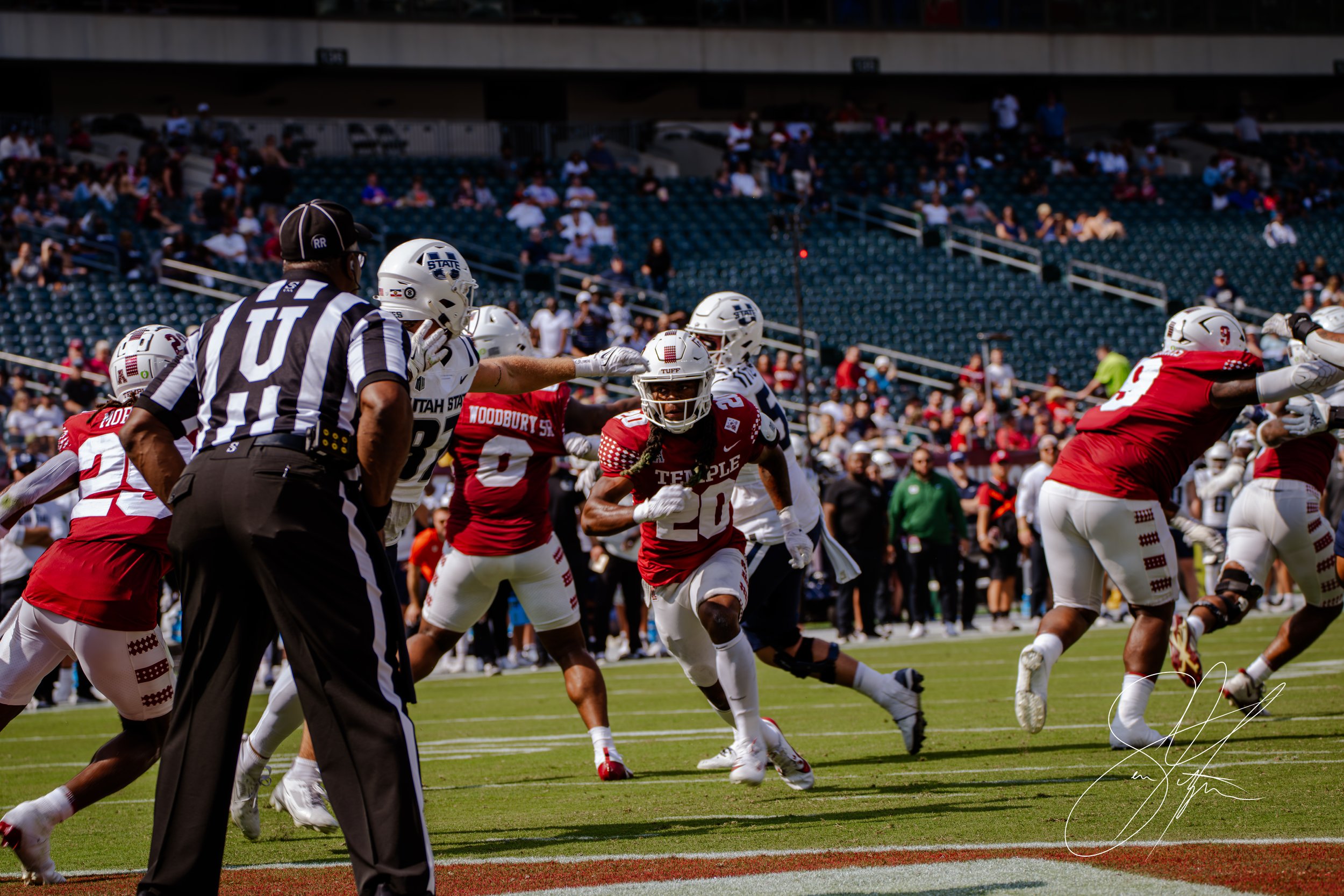 College football game with Temple player running with ball, Utah State players defending, referee in foreground.