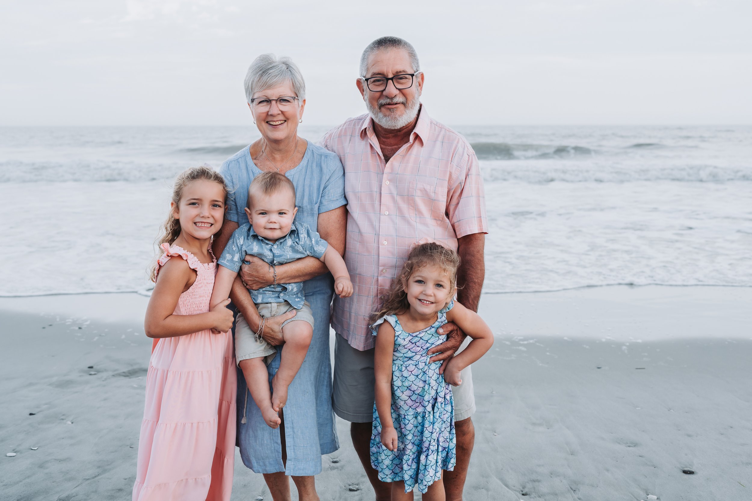 A family of five, including two older adults and three children, smiling on a beach with the ocean in the background.