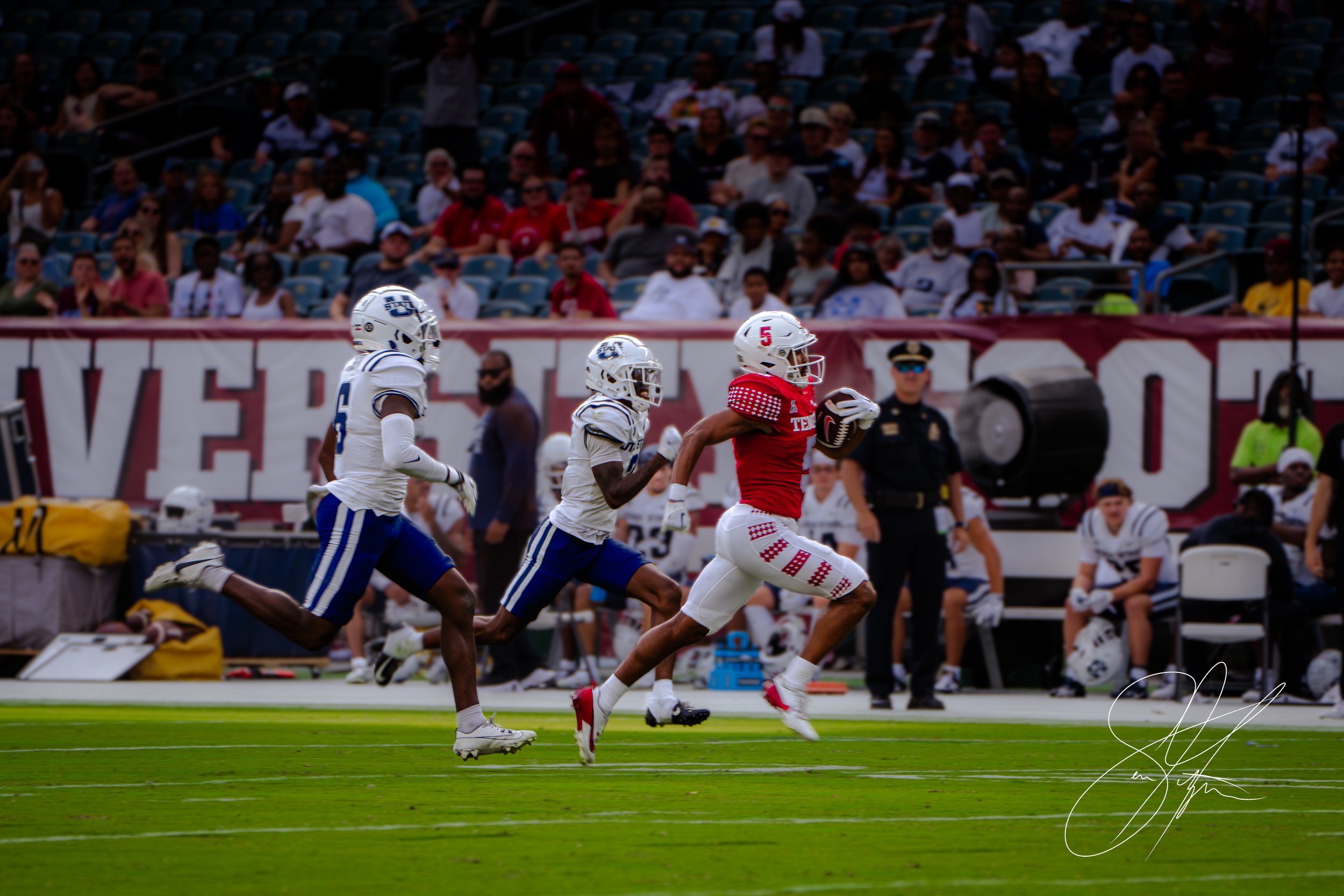 Football player in red uniform running with the ball pursued by two players in white uniforms on a field during a game.