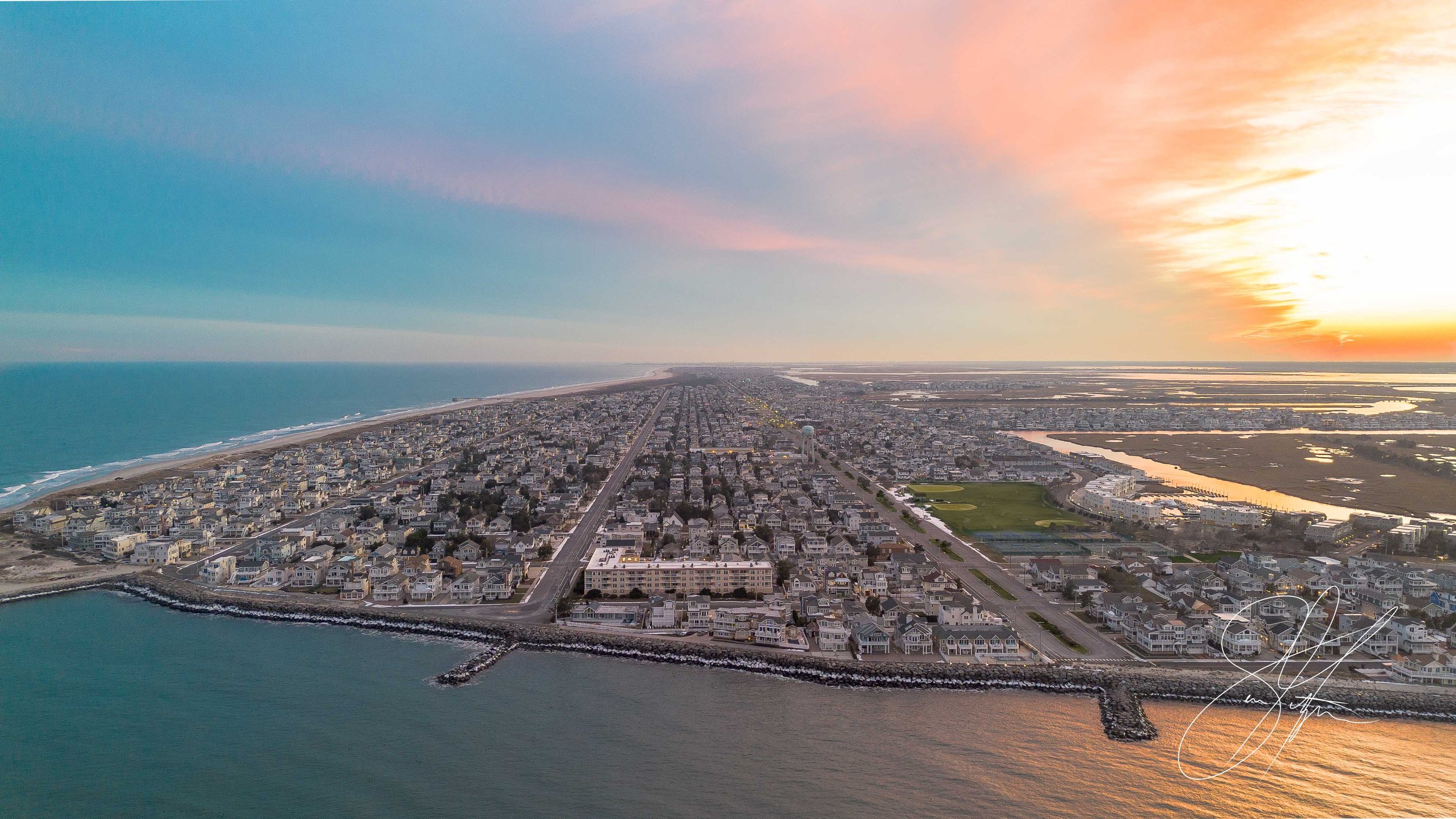 Aerial view of a coastal city with residential houses along the shoreline and a sunset over the ocean, creating a colorful sky.