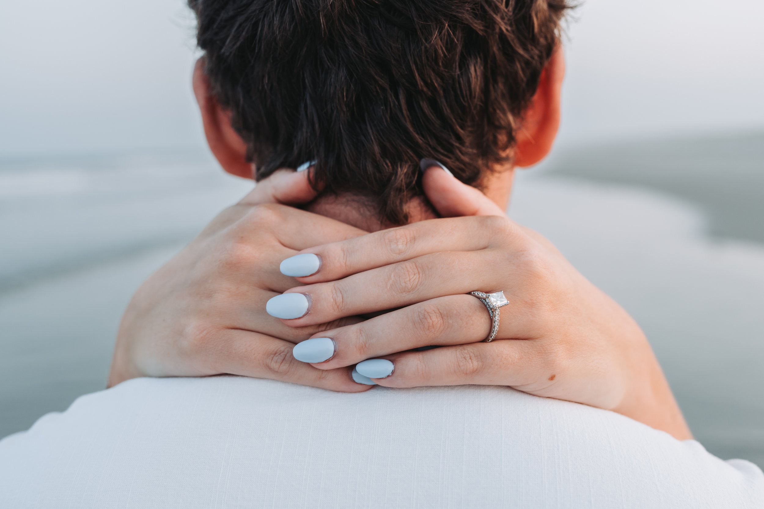 Person with blue-painted nails and an engagement ring embracing another individual from behind, beach background.