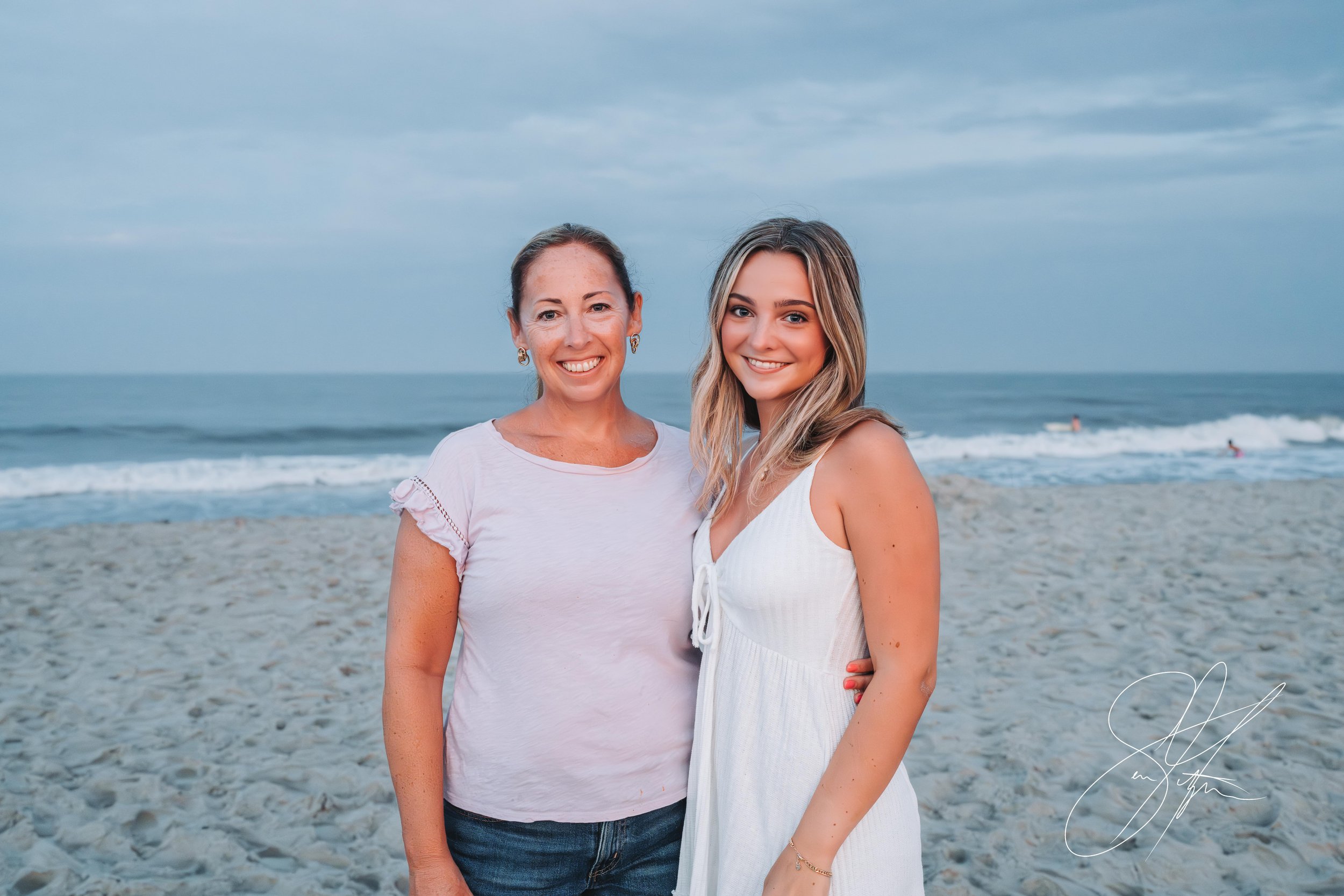 Two women smiling on a beach with ocean waves in the background, overcast sky, wearing casual summer outfits.