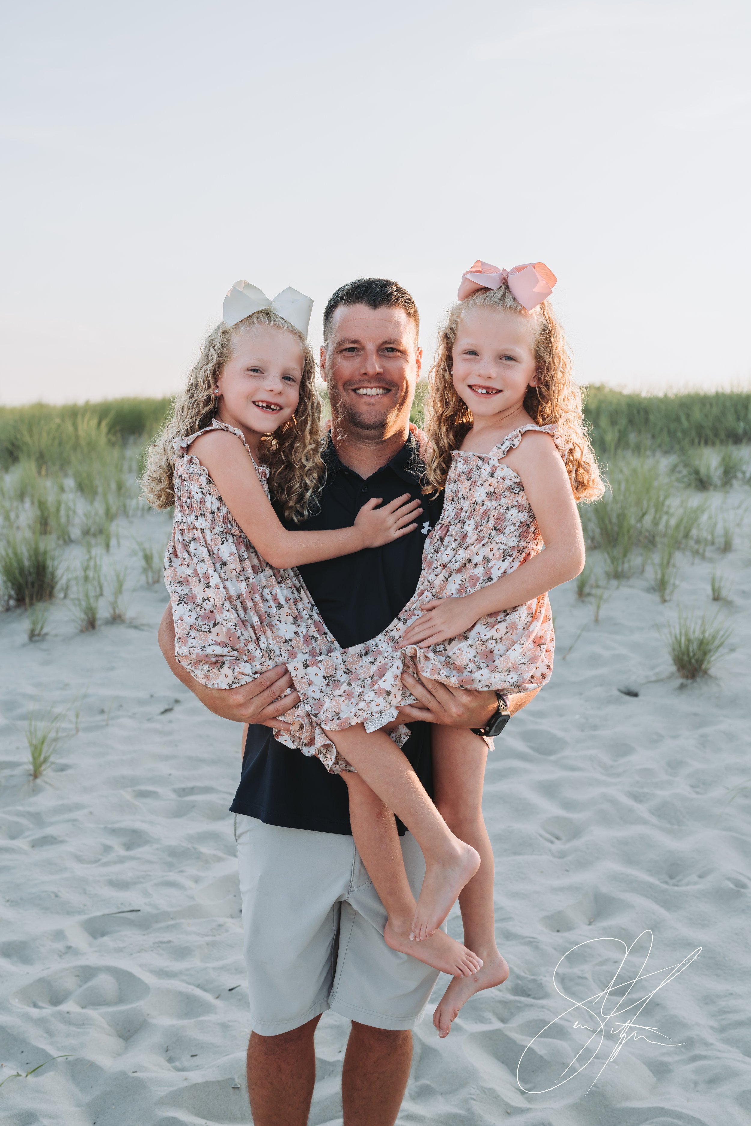 A man holding two young girls on a sandy beach, both wearing similar floral dresses and large bows in their hair.