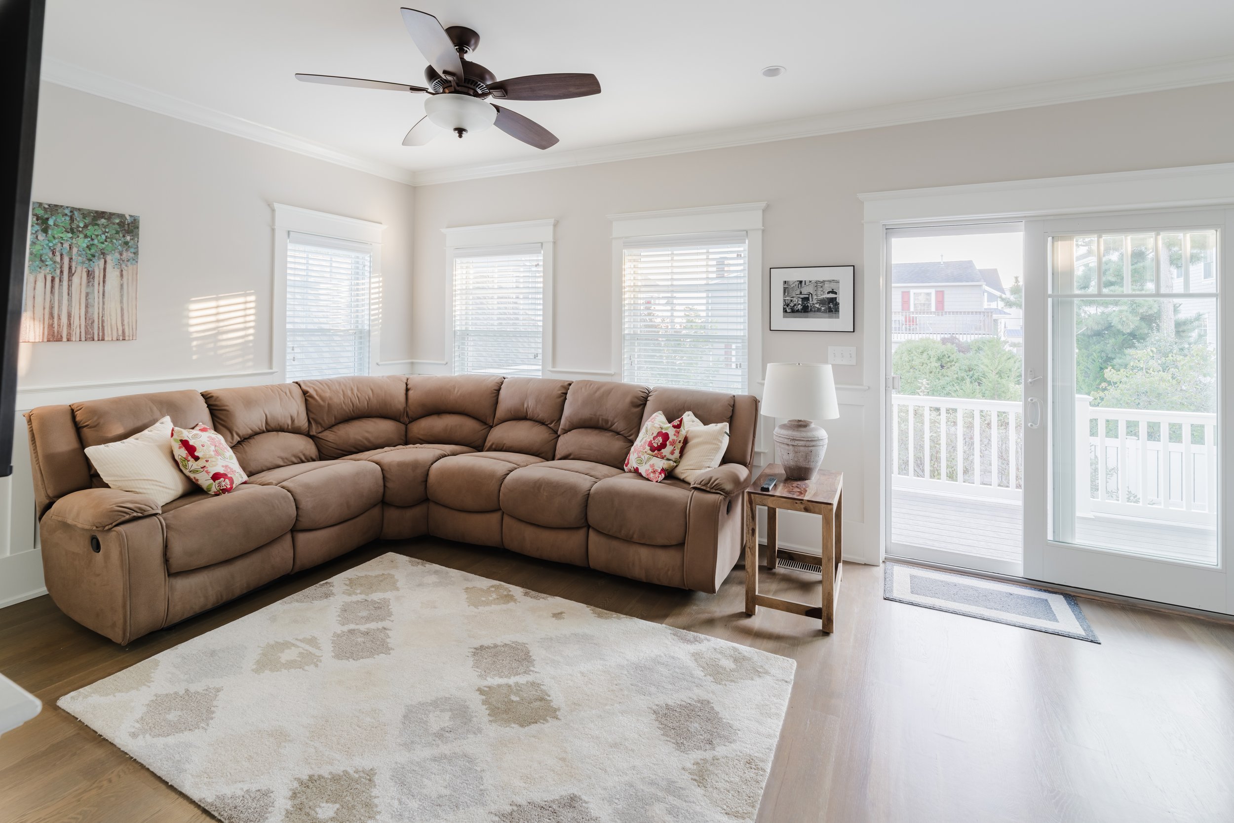 Cozy living room with a large beige sectional sofa, decorative pillows, a wooden side table with a lamp, a patterned area rug, and multiple windows providing natural light. A ceiling fan is installed above, and a sliding glass door leads to a balcony