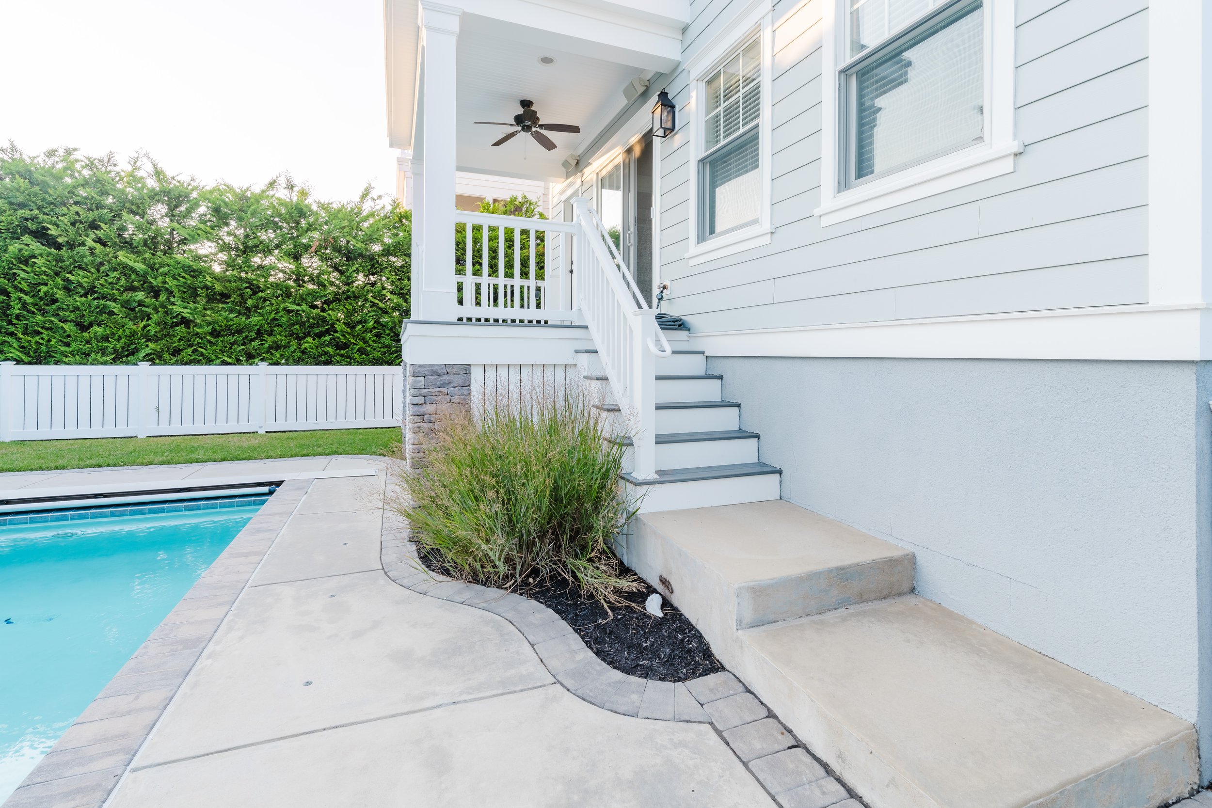 Outdoor scene with a light blue house, white stairs and railing, a porch with a ceiling fan, patio with a swimming pool, and a fence with hedges.
