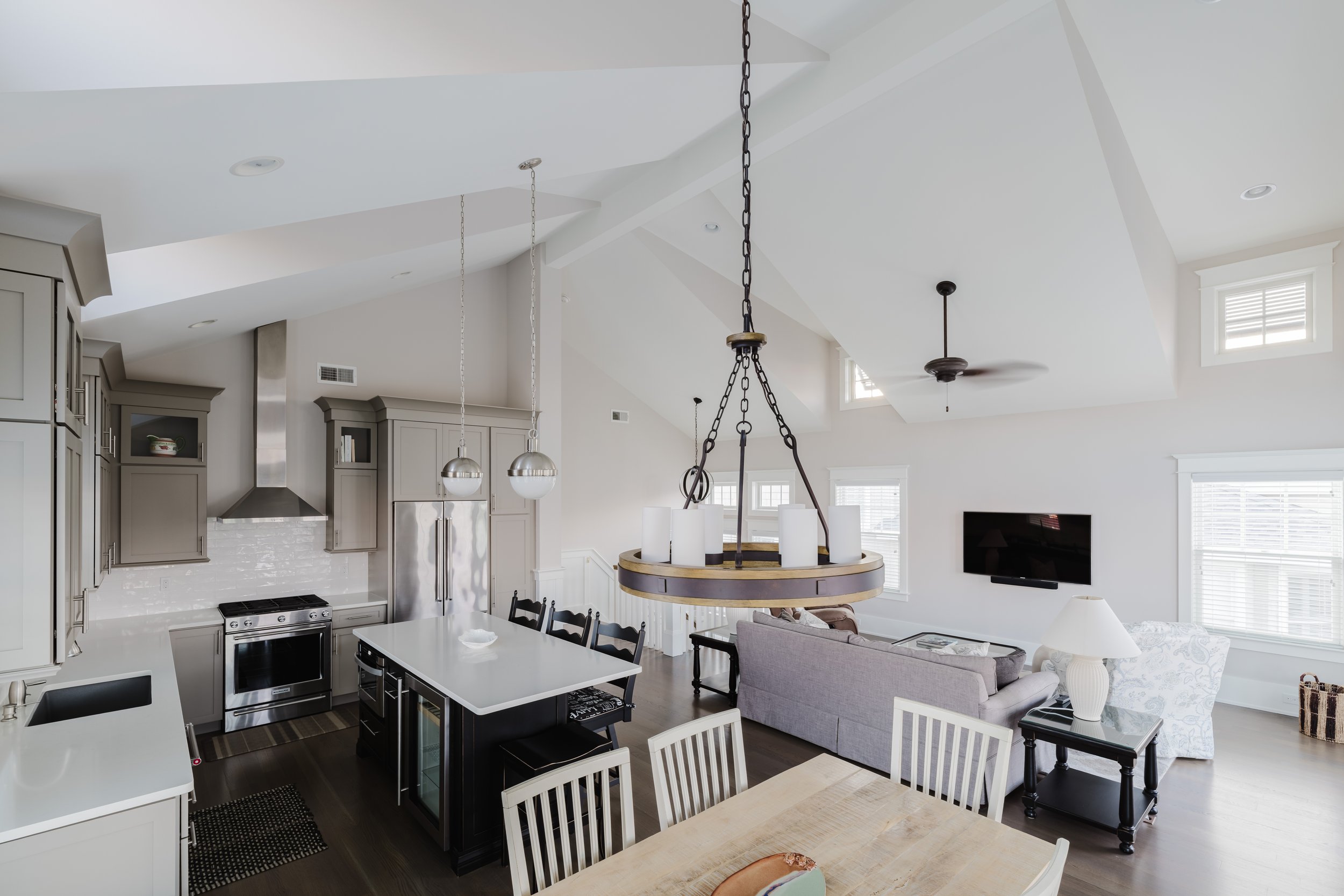 Open-concept kitchen and living room with modern design; gray cabinetry, stainless steel appliances, island with white countertop, dining table with chairs, sofa, TV, ceiling beams, pendant lights, and windows.