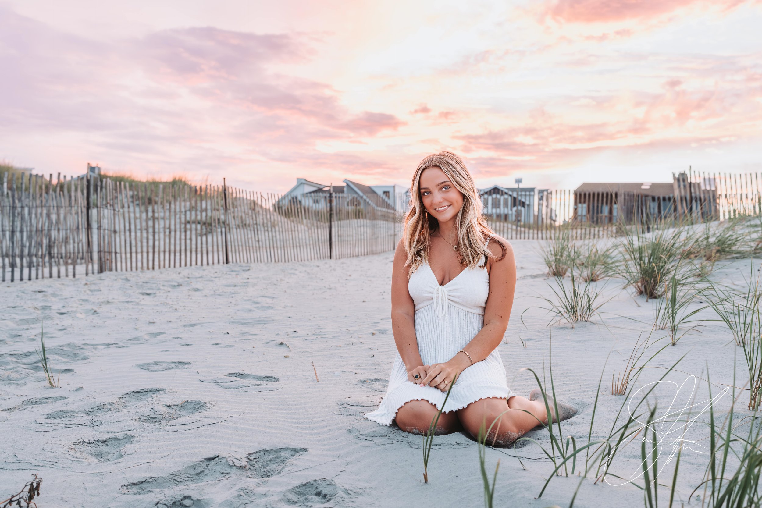 Woman in white dress kneeling on sandy beach with beach grass, fence, and houses in the background at sunset.