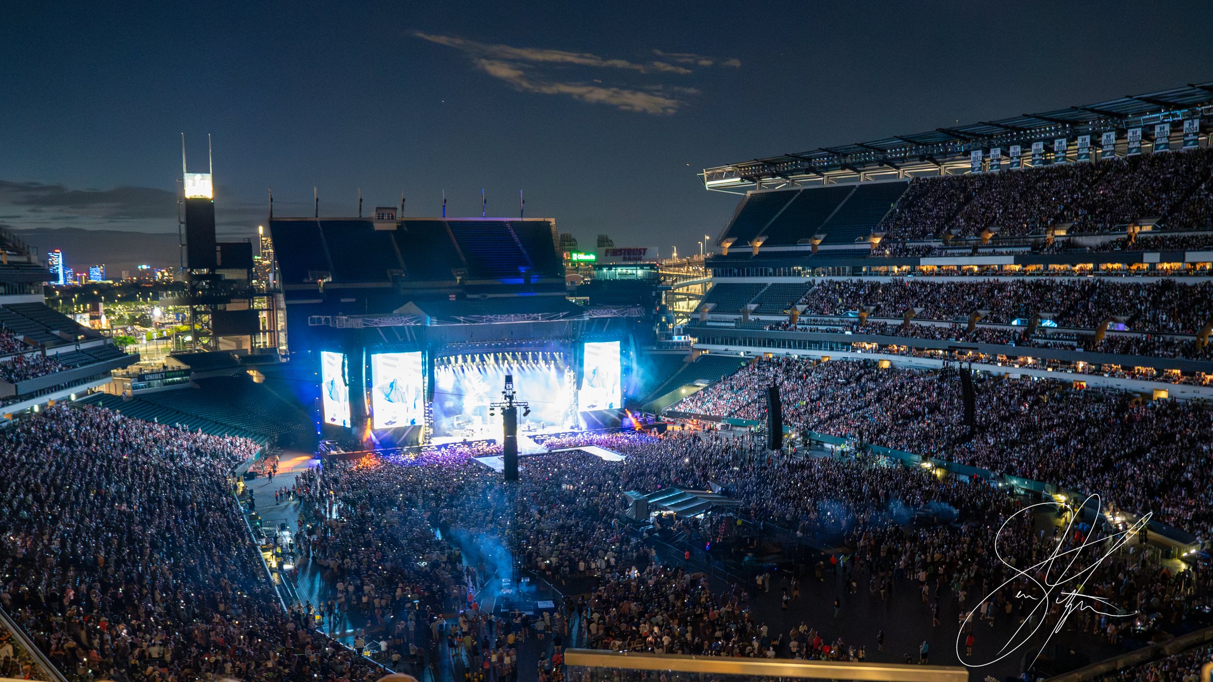 A large outdoor concert at a stadium with a full audience at night, illuminated stage with large screens, city lights in the background.