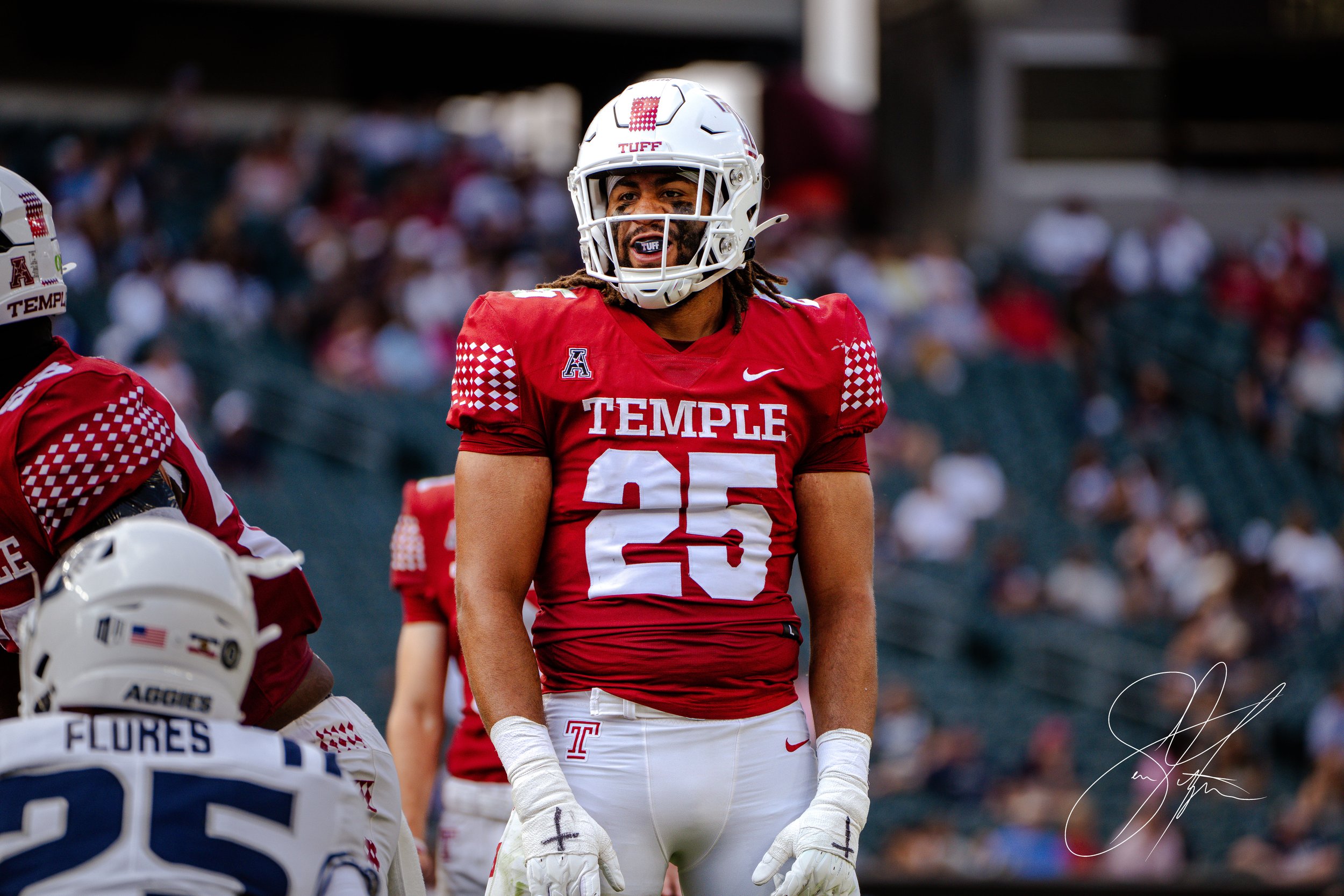 A football player in a red Temple University jersey, number 25, stands on the field during a game. He wears a helmet and white gloves. The crowd is blurred in the background.