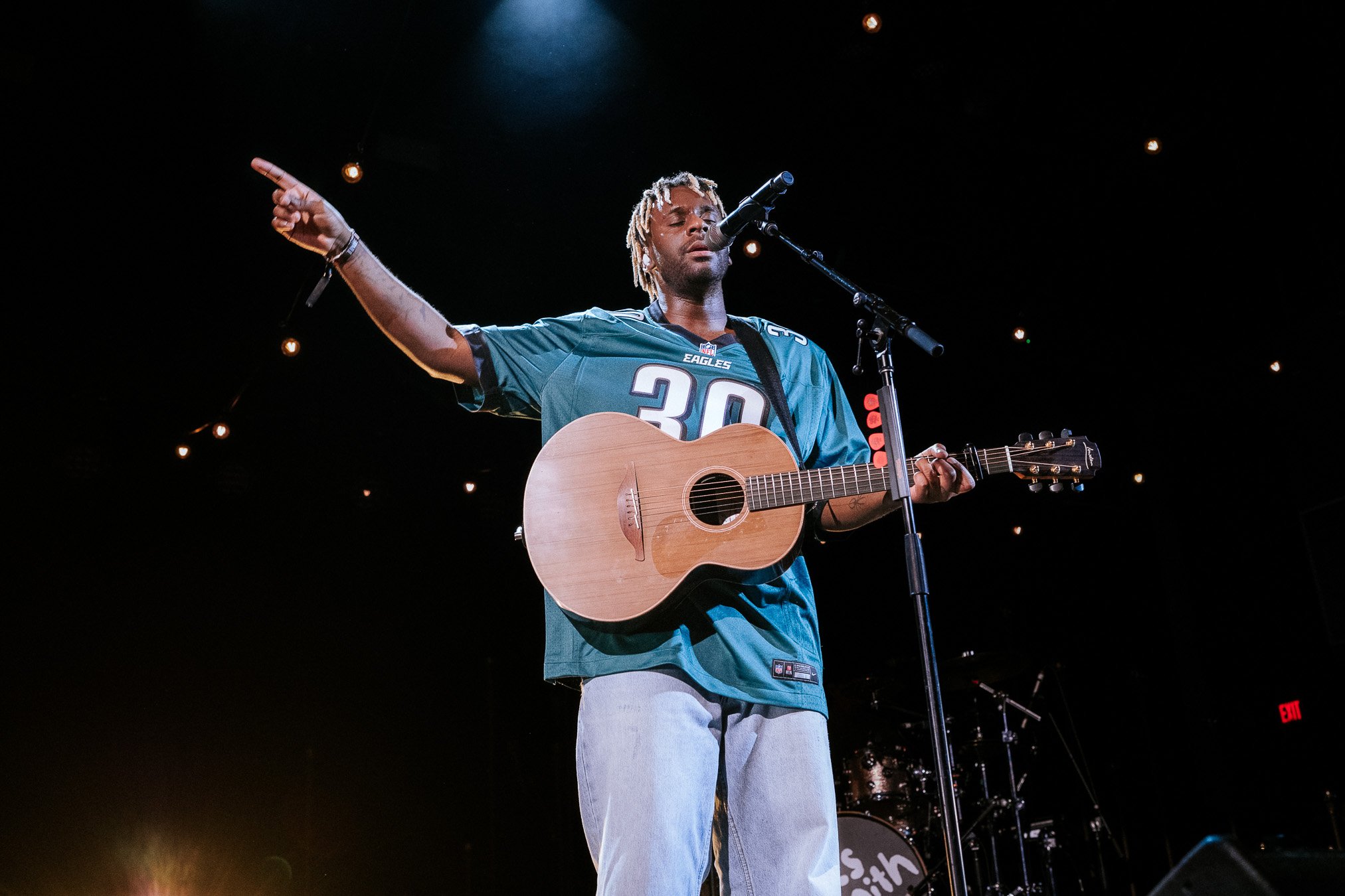 Musician wearing a green football jersey with number 30, playing acoustic guitar, pointing, and singing on stage with microphone and dim lighting.