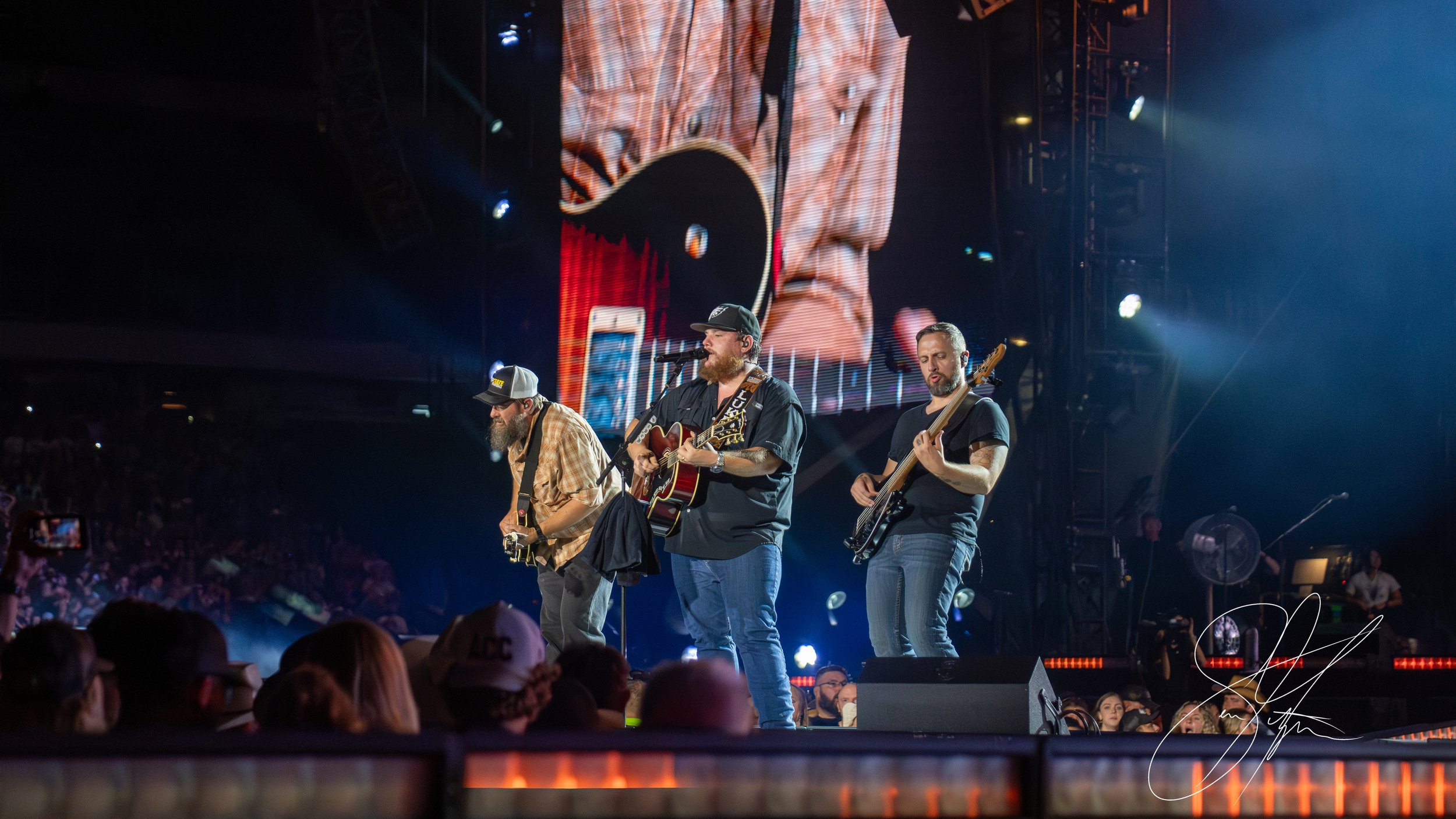 Three musicians performing on stage with guitars, large screen displaying a close-up, crowd watching in the foreground.