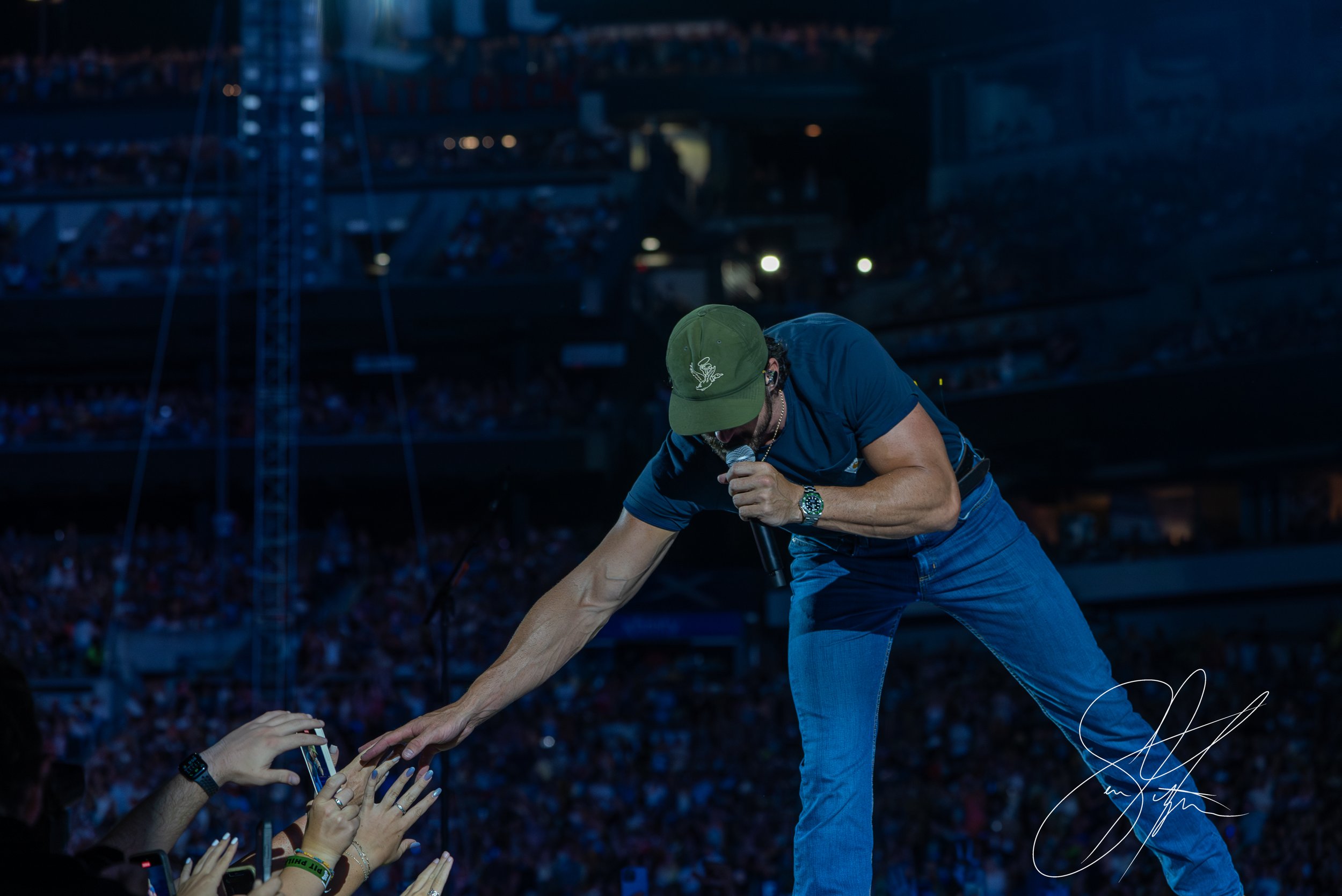 Man performing on stage, reaching out to crowd, stadium background.