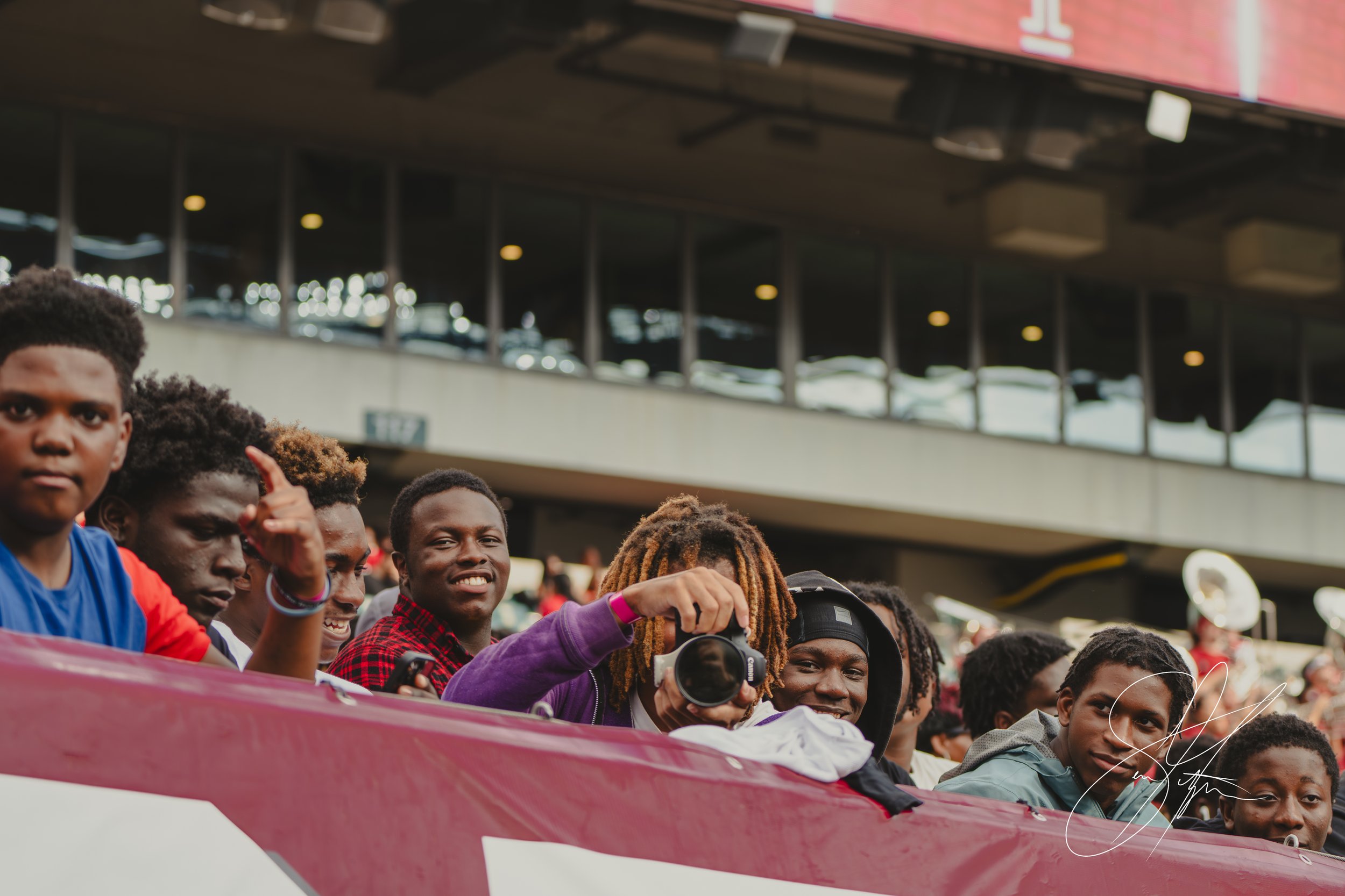 Group of young people smiling and taking photos at a sports event, with a stadium in the background.