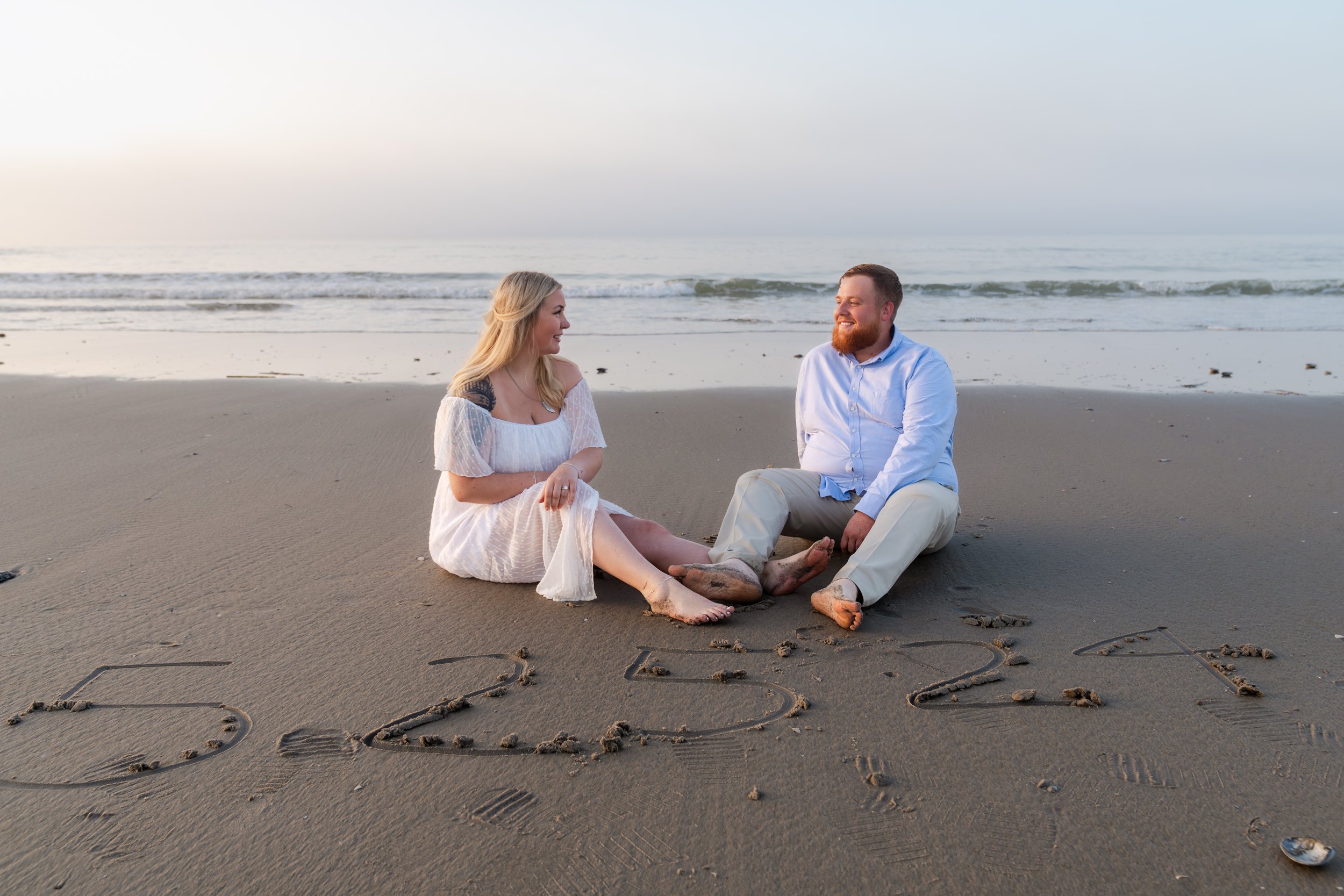 A couple sitting on a beach with a date "5-25-24" written in the sand, with ocean waves in the background.