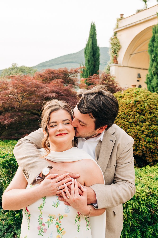 A man kisses a woman on her cheek as they hug in a garden with bushes, trees, and a building in the background.