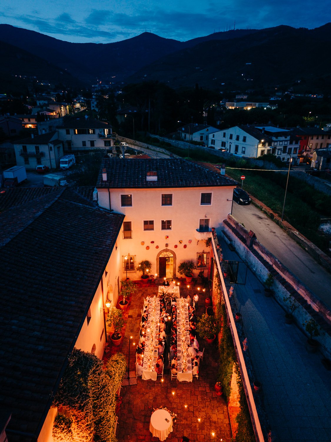 An aerial view of an outdoor dinner party at dusk in a courtyard, illuminated by string lights, with a large table filled with people, surrounded by potted plants and walls of a building, with a mountainous landscape in the background.