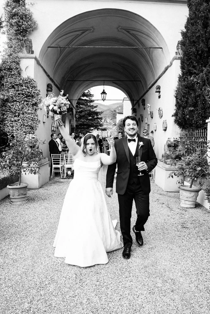 A bride and groom celebrating their wedding under an archway with guests seated in the background. The bride is holding a bouquet and raising her arms, while the groom is holding a glass and smiling.