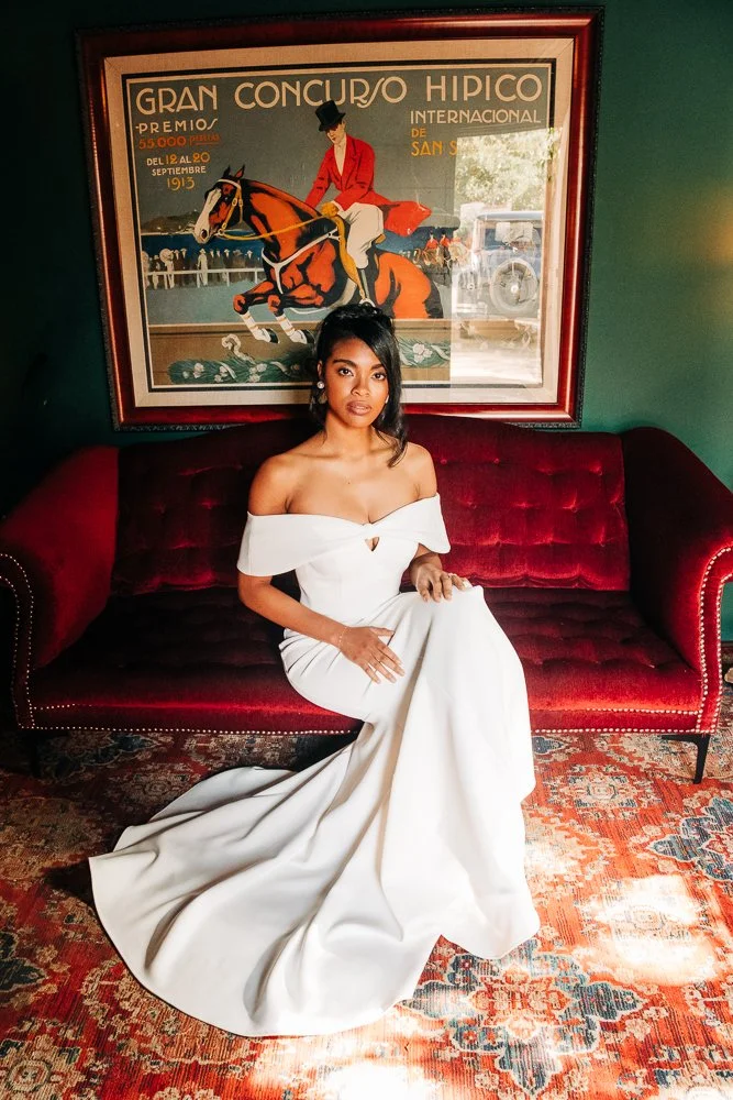 bridal portrait of a woman wearing a tailored white wedding dress, seated on a red velvet couch in a dark green room