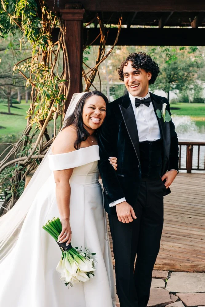 A couple laughs together, outdoors by a lake surrounded by trees, minutes after their wedding ceremony