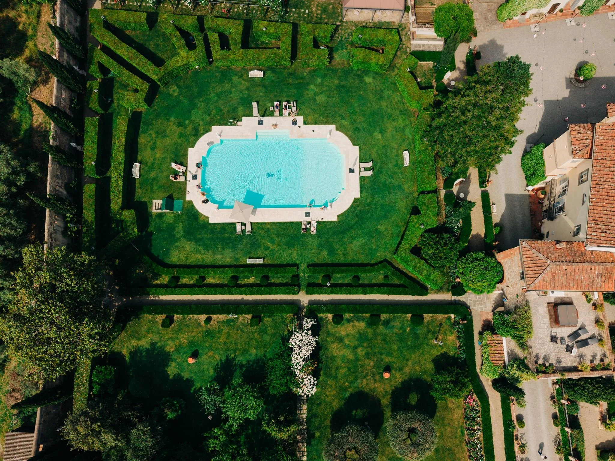 An aerial view of a backyard with a swimming pool, surrounded by a lush green lawn, hedges, and various trees. There are lounge chairs, umbrellas, and some people enjoying the area. The backyard is enclosed by a fence and neighboring houses are visible on one side.