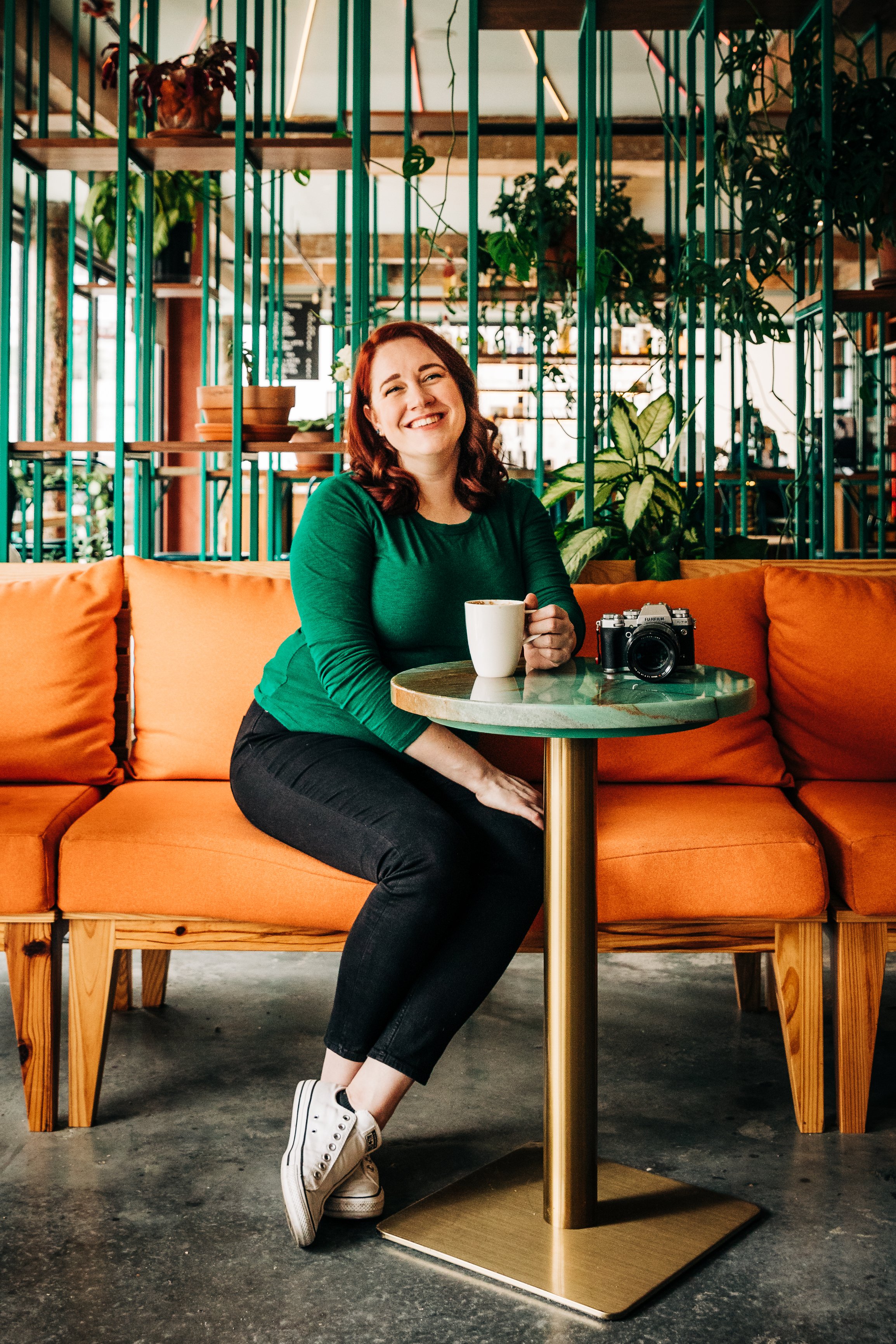 A woman with red hair smiling and sitting on an orange sofa at a small round table in a cozy, well-lit cafe filled with potted plants and greenery. She is holding a white mug, with a camera placed on the table near her. She is wearing a green top, black jeans, and white sneakers.