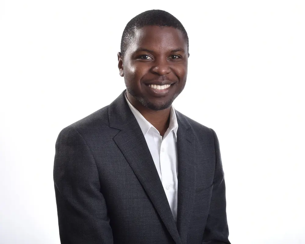 Portrait of a smiling Black man in a dark suit and white shirt against a white background.