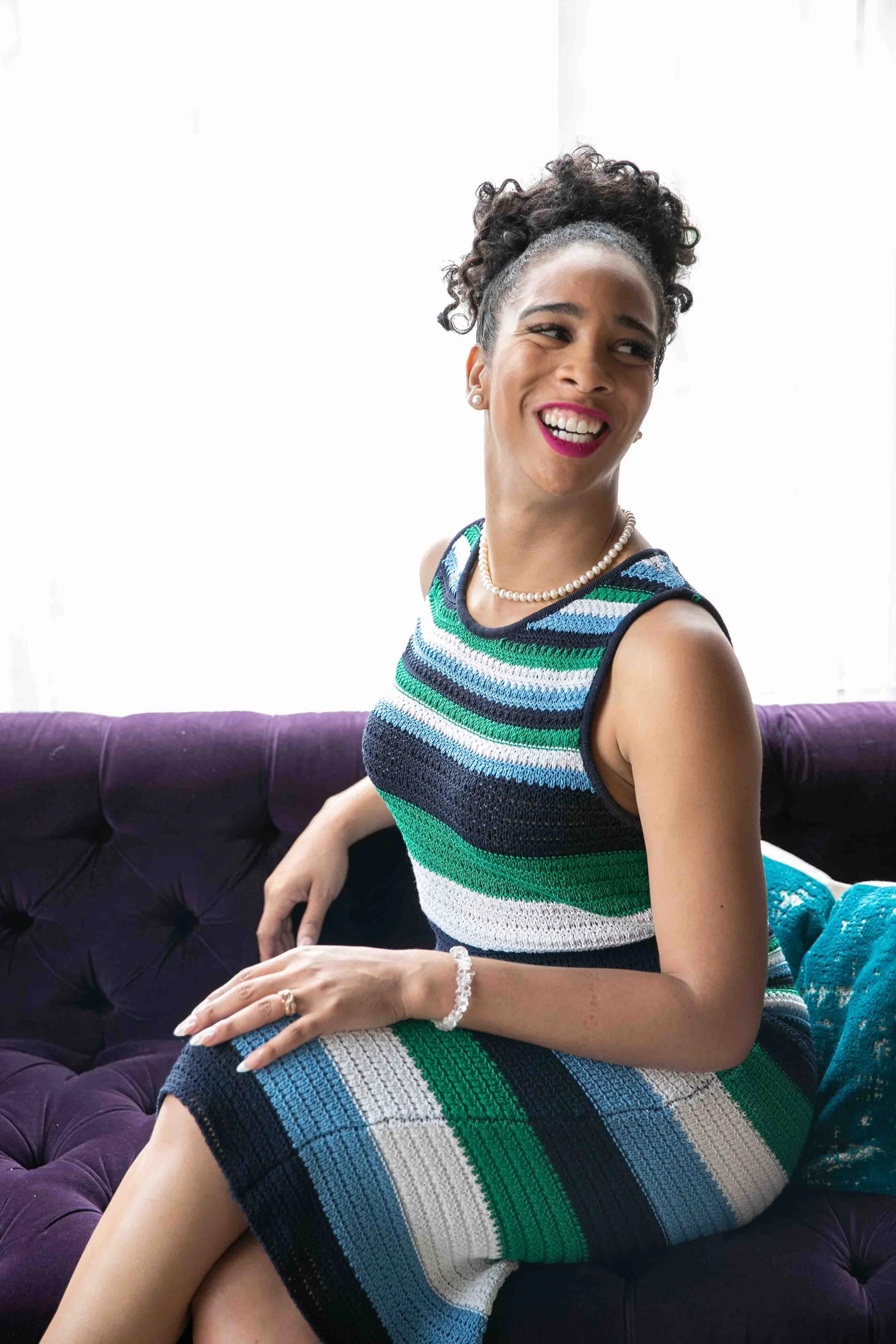 A woman smiling and sitting on a purple sofa, wearing a colorful striped sleeveless dress, pearl necklace, bracelet, and earrings.