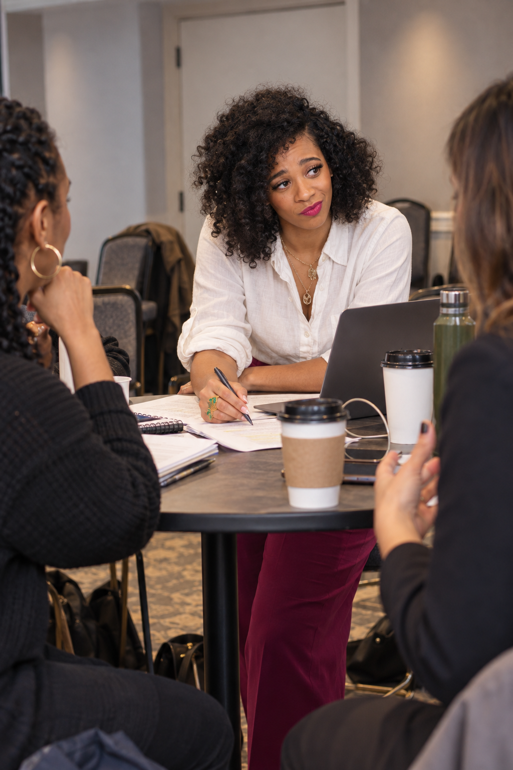 A woman with curly hair wearing a white blouse is sitting at a table, speaking to two women across from her. The table has coffee cups, a laptop, a green water bottle, and notebooks.