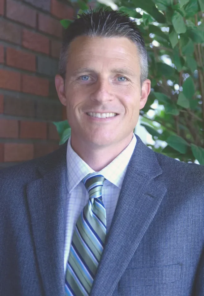 A man in a suit and tie standing outdoors next to a brick wall and green foliage, smiling at the camera.