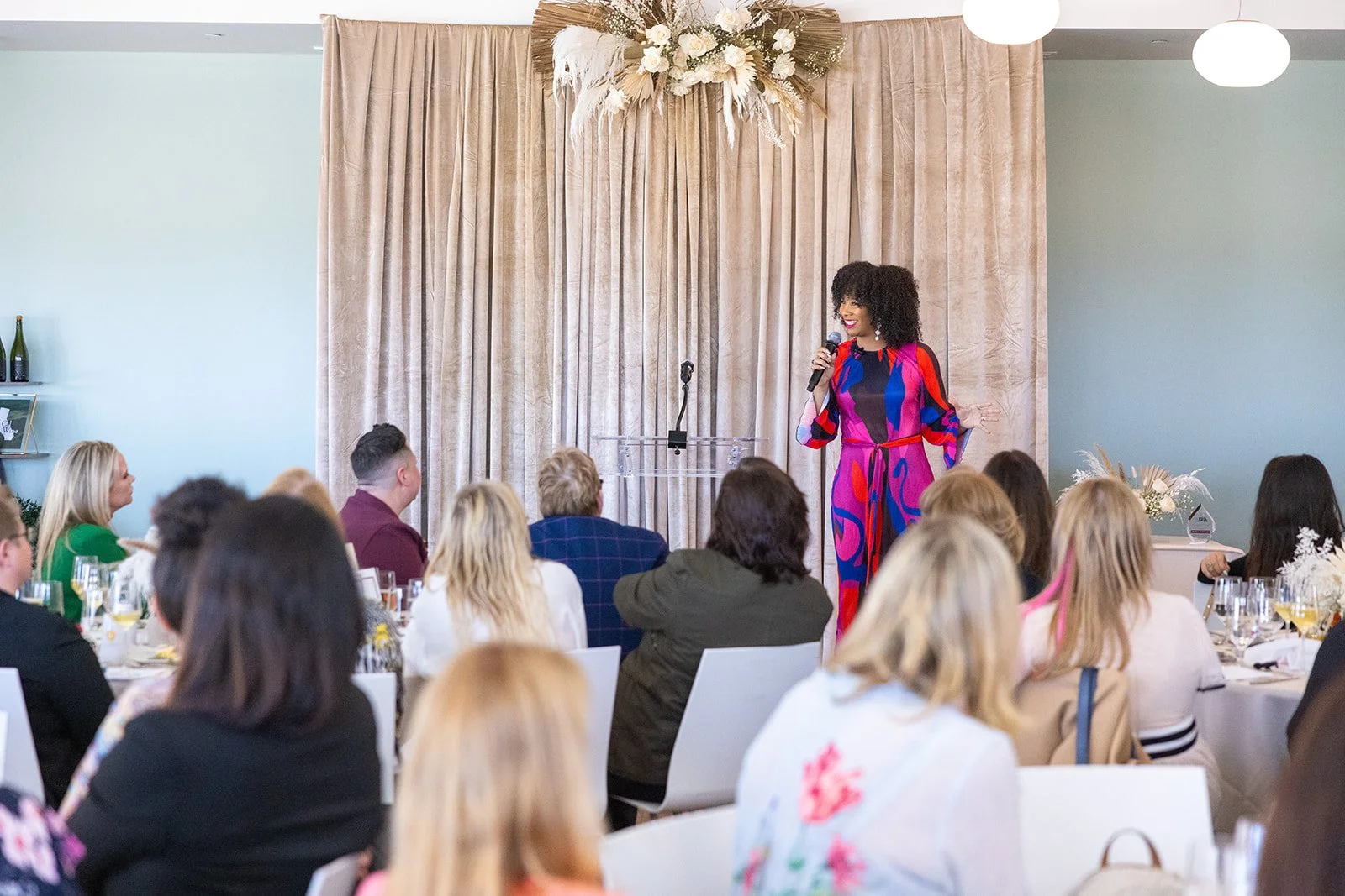 A woman standing on stage speaking into a microphone at a formal event, with an audience seated at tables in front of her. The stage has beige curtains and floral decorations.