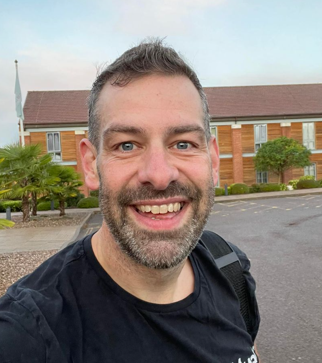 A man with a beard smiling outdoors, wearing a black shirt, with a building and trees in the background.