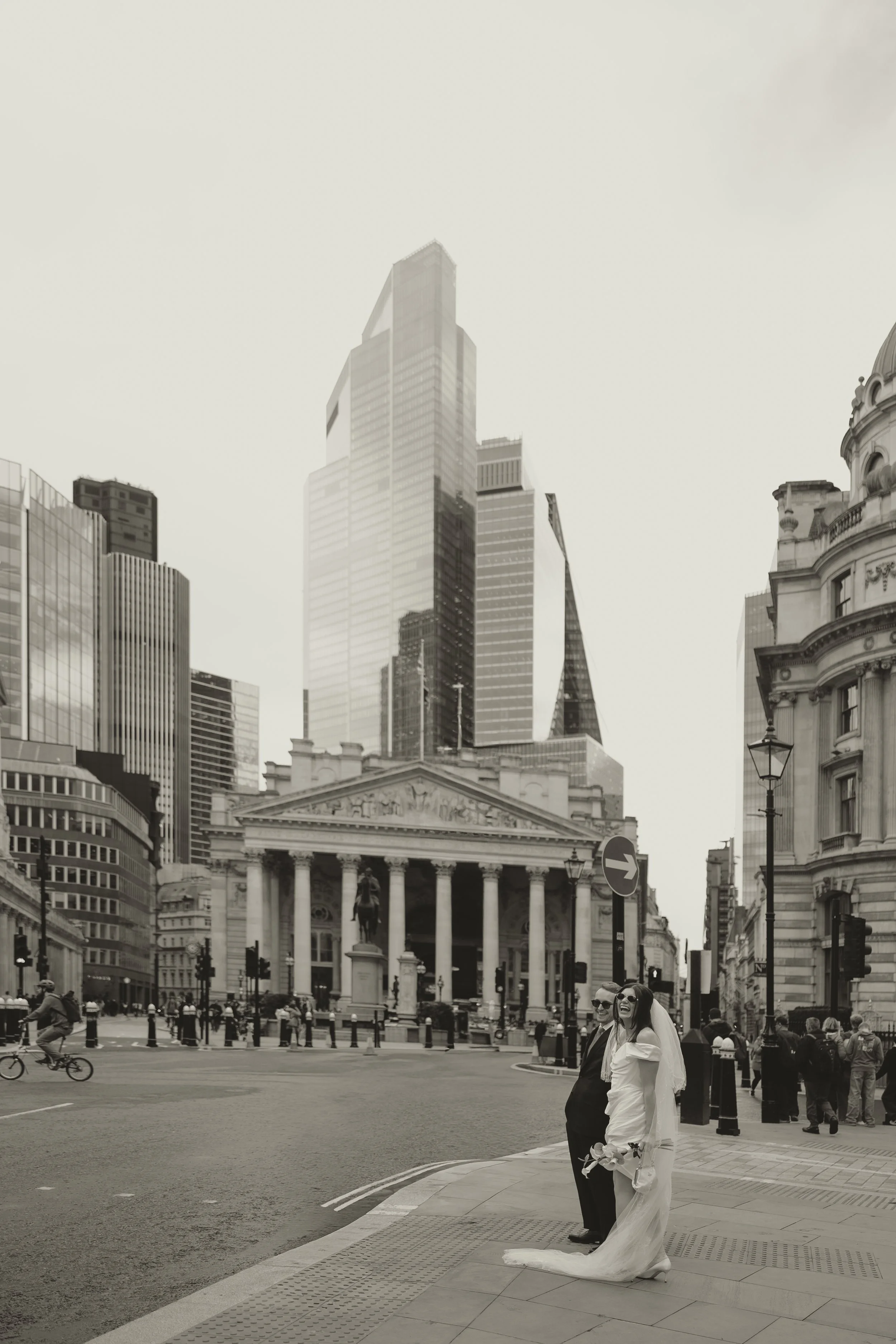 Editorial wedding photography - a bride and groom standing on a city street with tall buildings and historic architecture in the background.