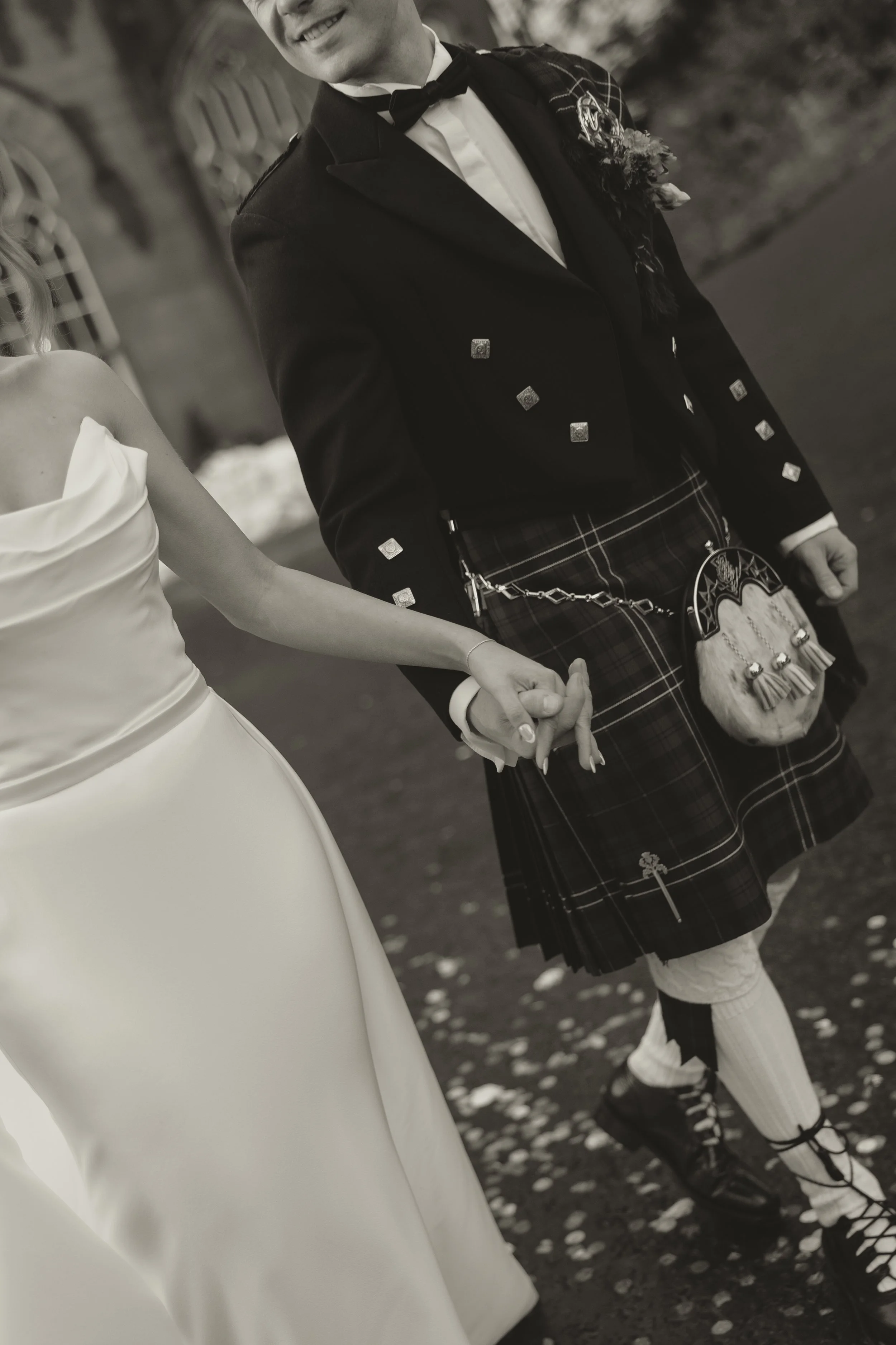 A bride and groom holding hands, dressed in wedding attire, with the bride in a white gown and the groom in traditional Scottish kilt and jacket, walking outdoors. Editorial wedding photo.